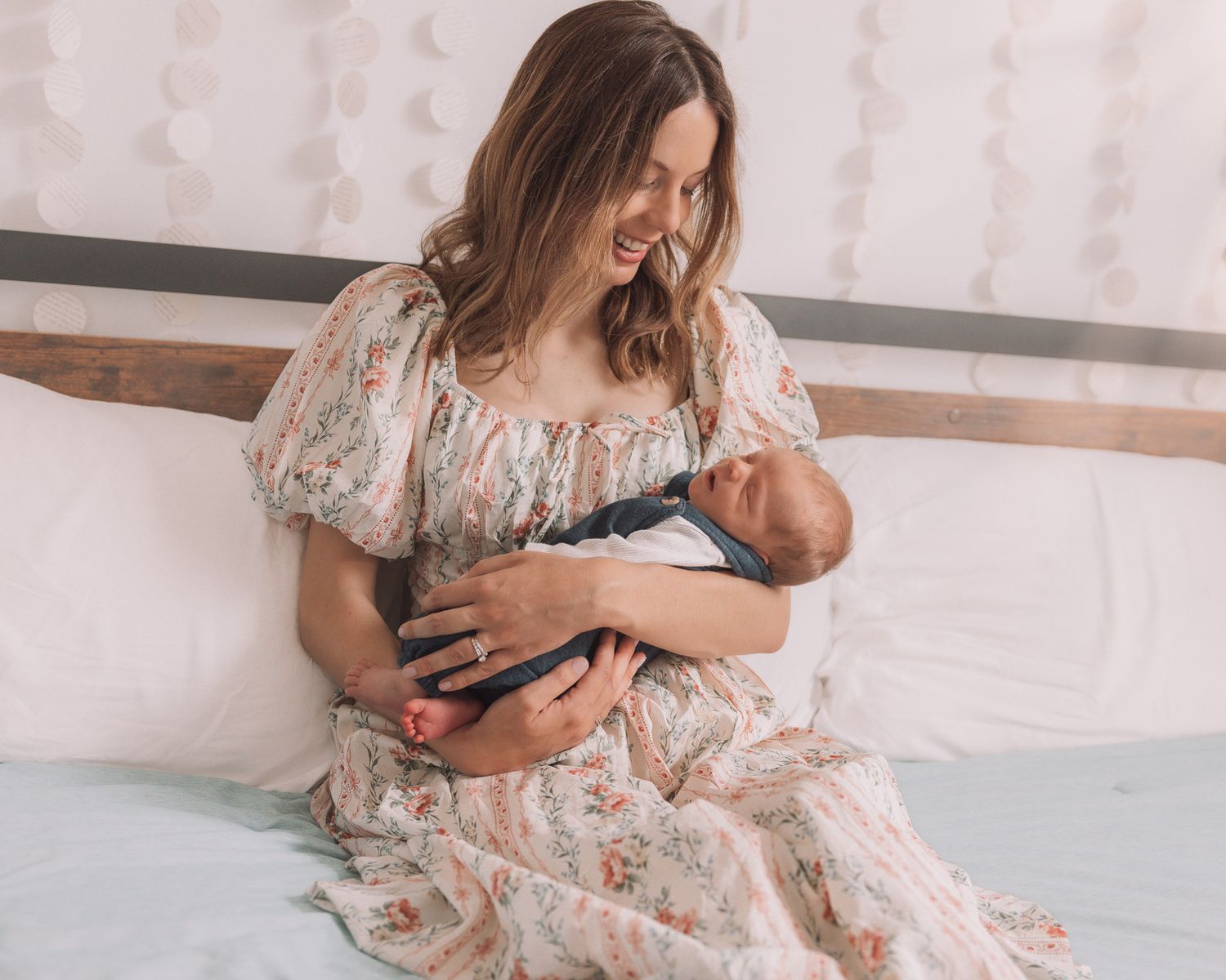 A smiling mother in a floral dress sits on a bed cradling her newborn baby.