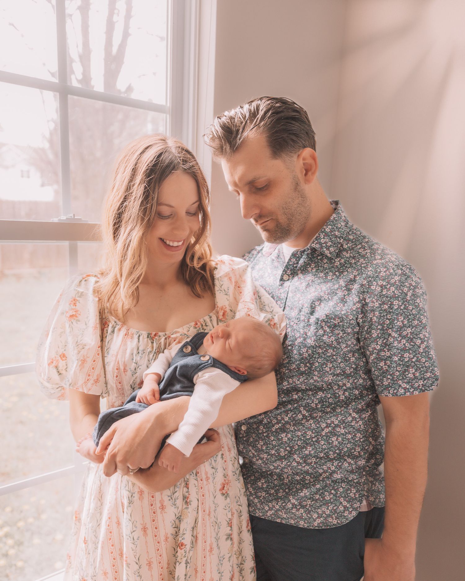 Happy parents in coordinating floral outfits holding their sleeping newborn baby by a sunlit window.