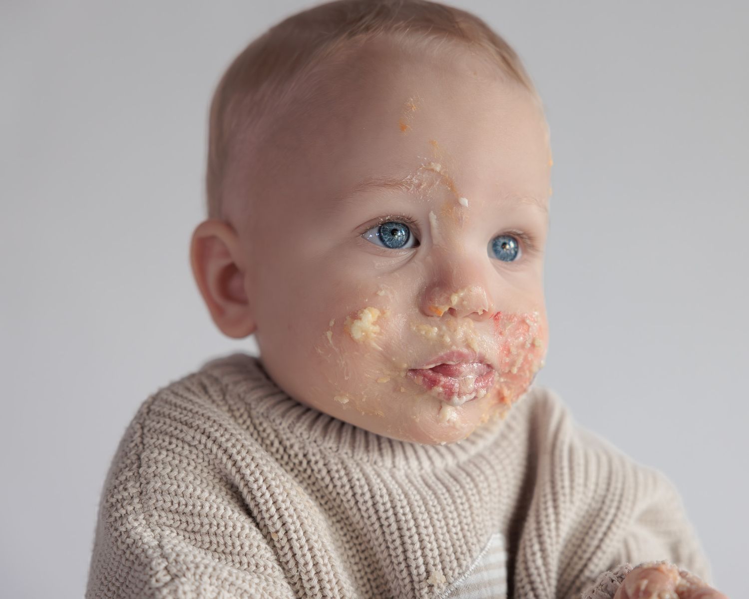 Close-up of blue-eyed baby with cake smeared on face wearing a cozy beige knit sweater.