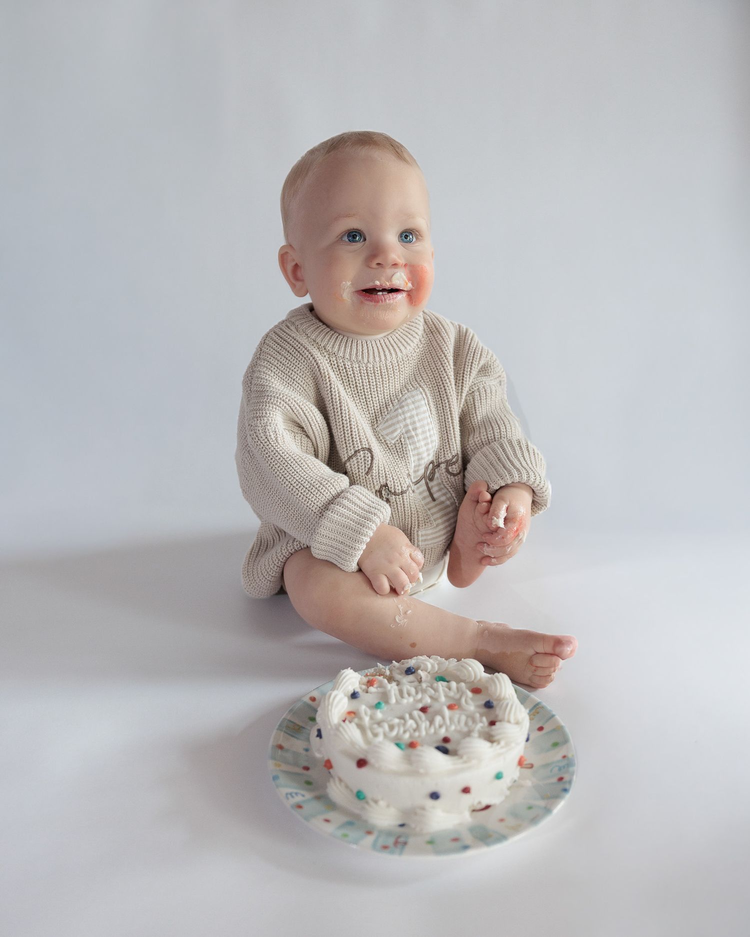 Smiling baby in beige knit romper sitting beside a white birthday smash cake with colorful sprinkles.