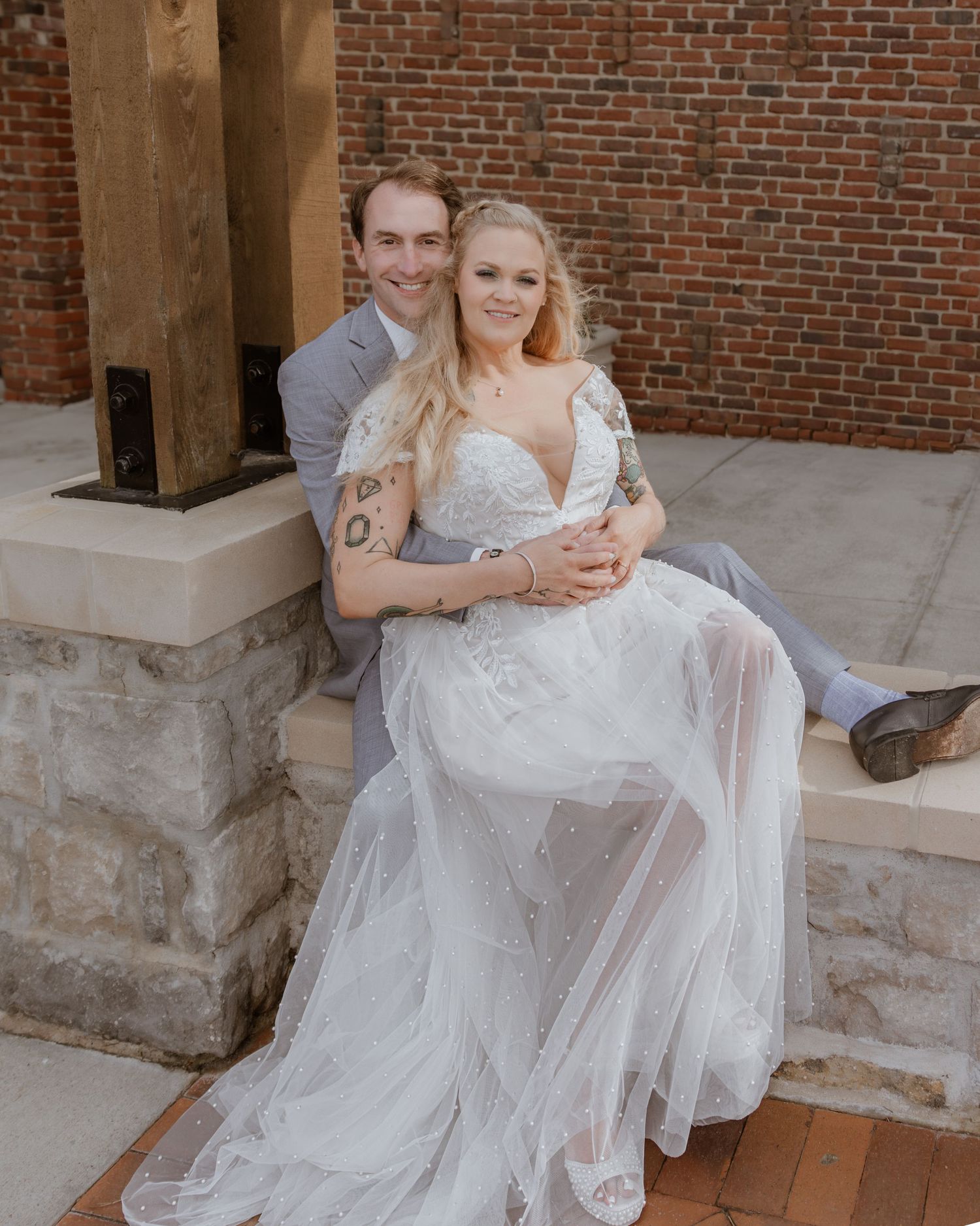 Bride in flowing tulle wedding dress and groom in gray suit embracing outdoors near brick wall.