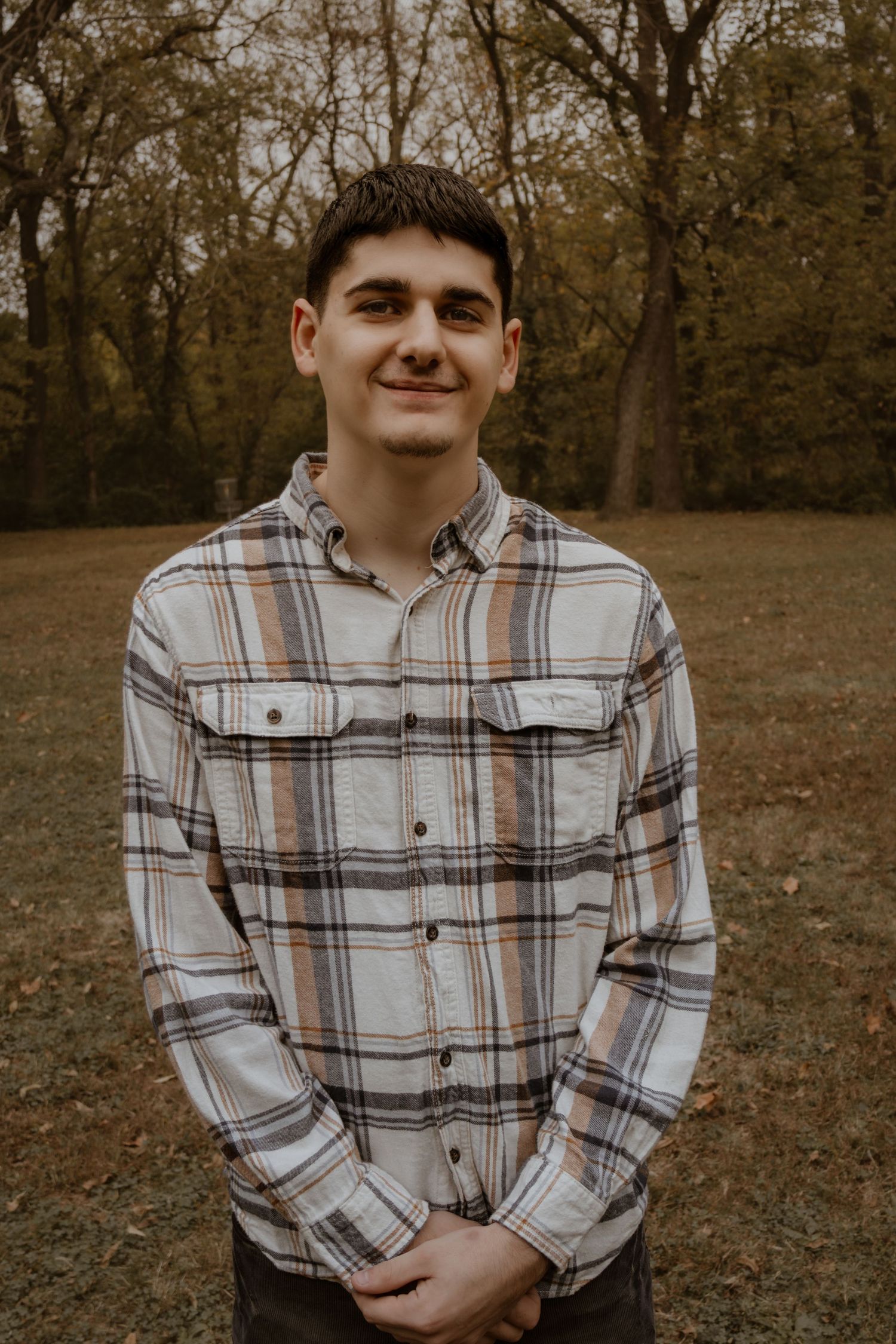 A young person in a plaid flannel shirt smiles at the camera in an autumn forest setting.