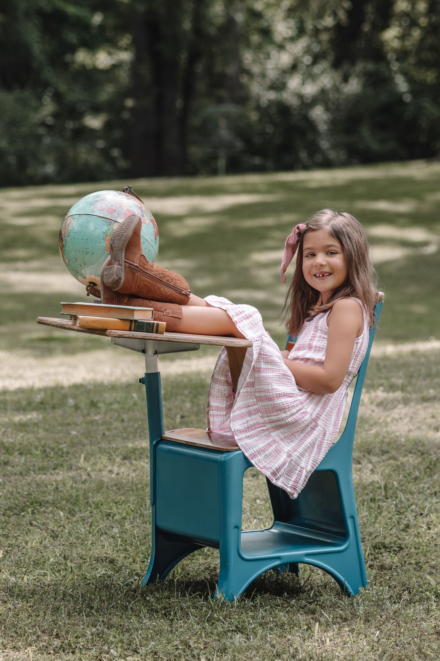 A young student sits at a vintage school desk outdoors in a grassy area on a sunny day.
