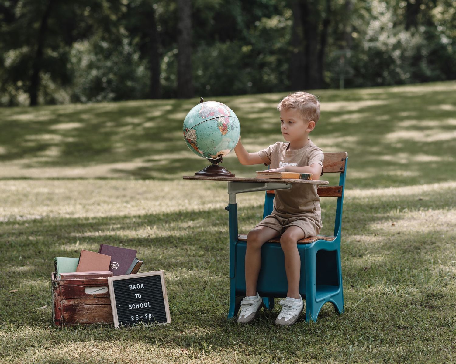 A child sits at a small turquoise table on a grassy lawn with a globe and chalkboard sign nearby.