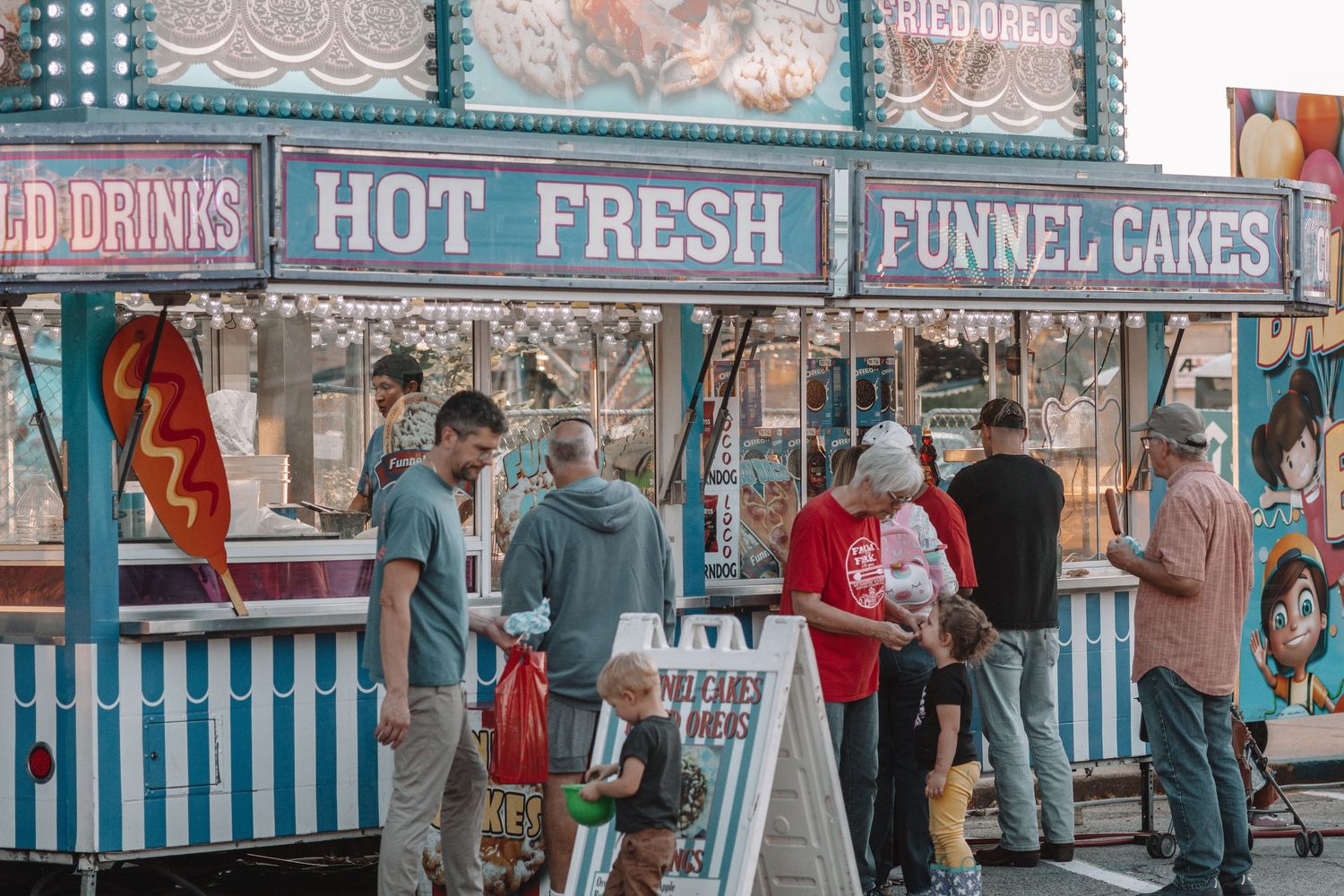Customers gather at a retro-style carnival concession stand with striped awnings selling cold drinks and funnel cakes.