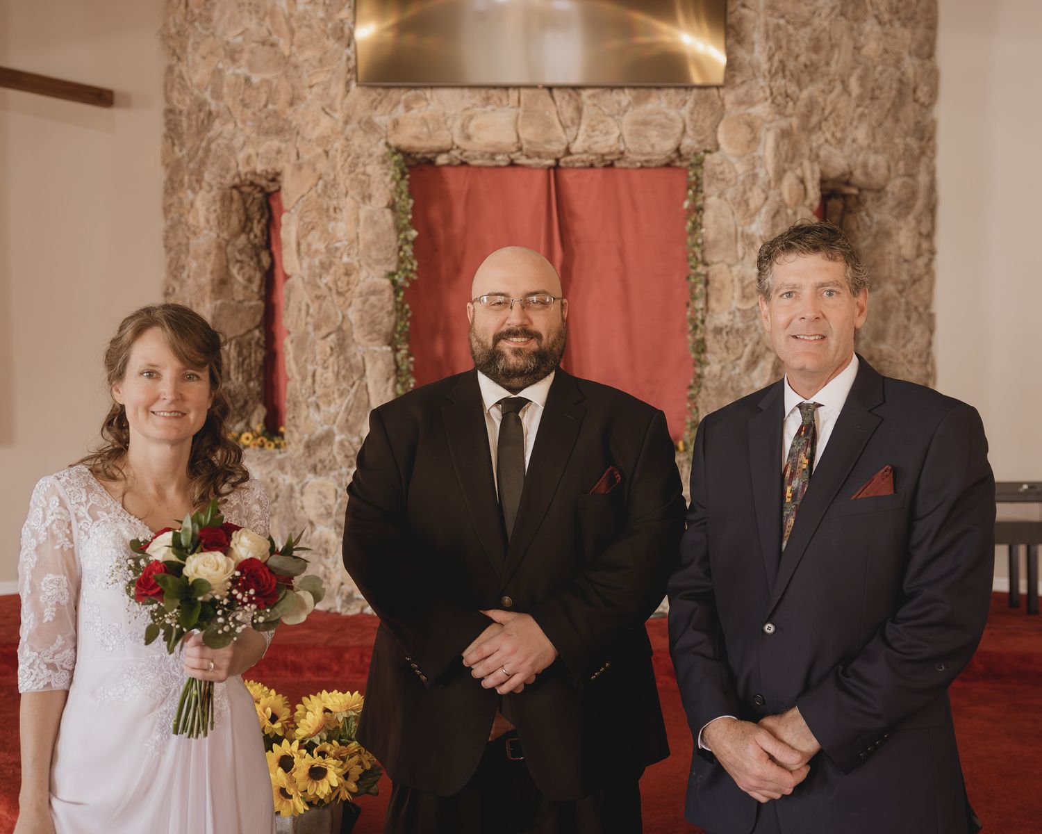 Bride holding floral bouquet stands with officiant and groom at elegant stone chapel wedding ceremony.