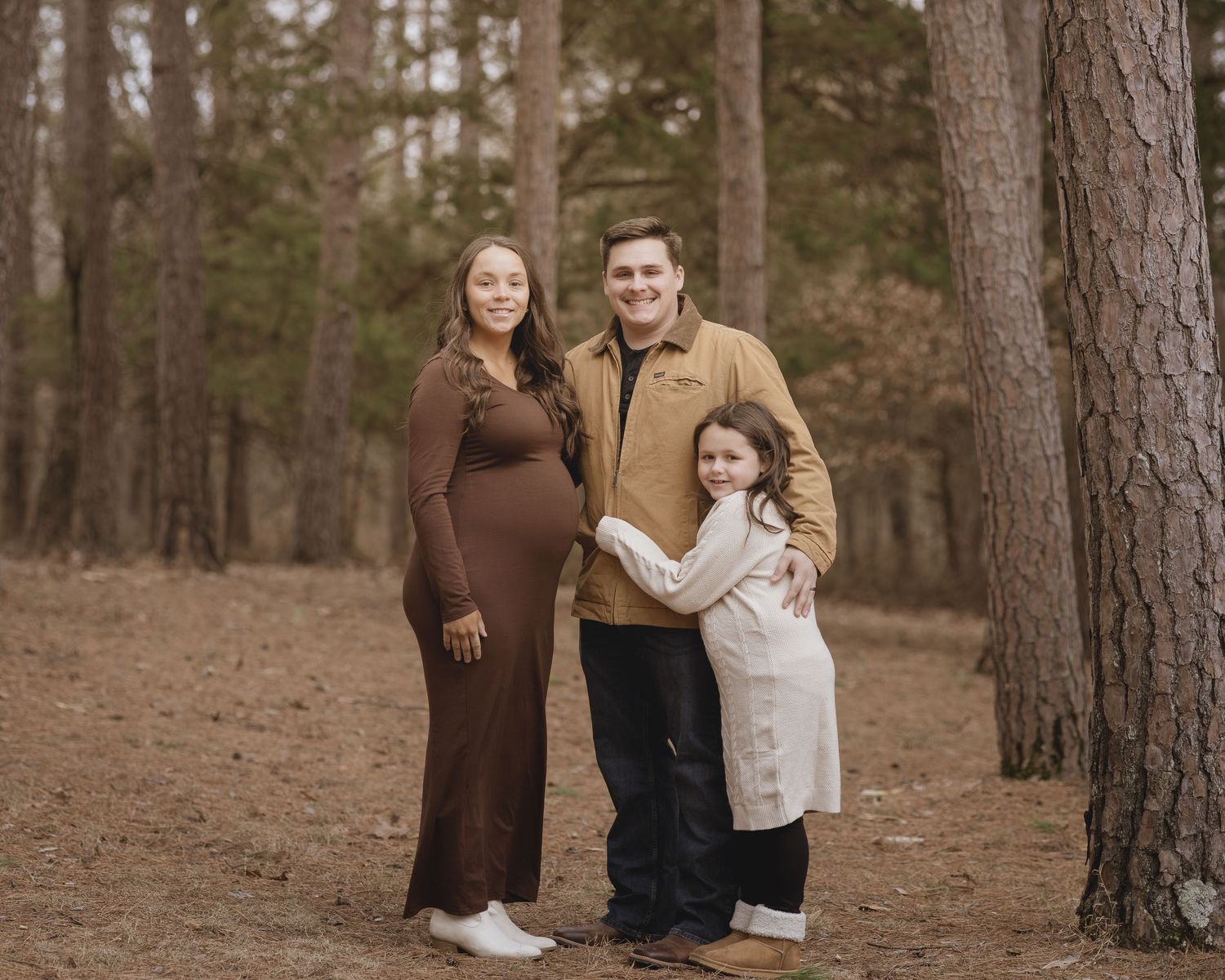 A couple poses together in a serene forest setting with tall pine trees and fallen leaves on the ground.