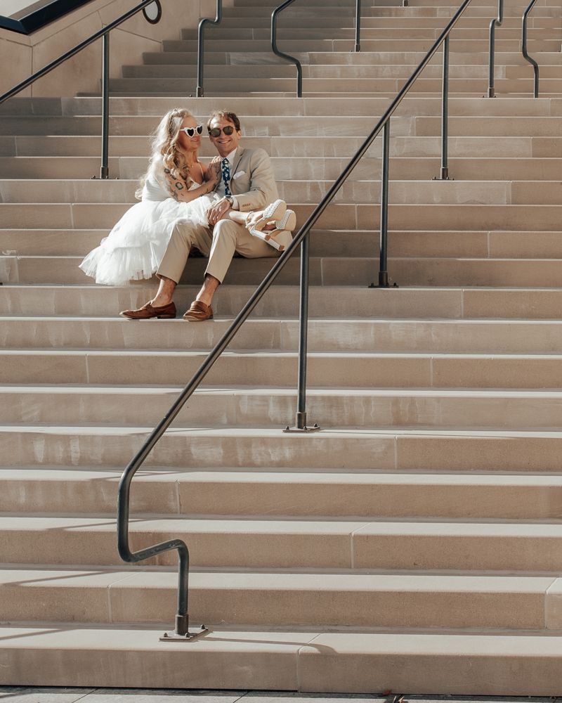 A couple sits together on outdoor concrete steps with a black metal railing on a sunny day.