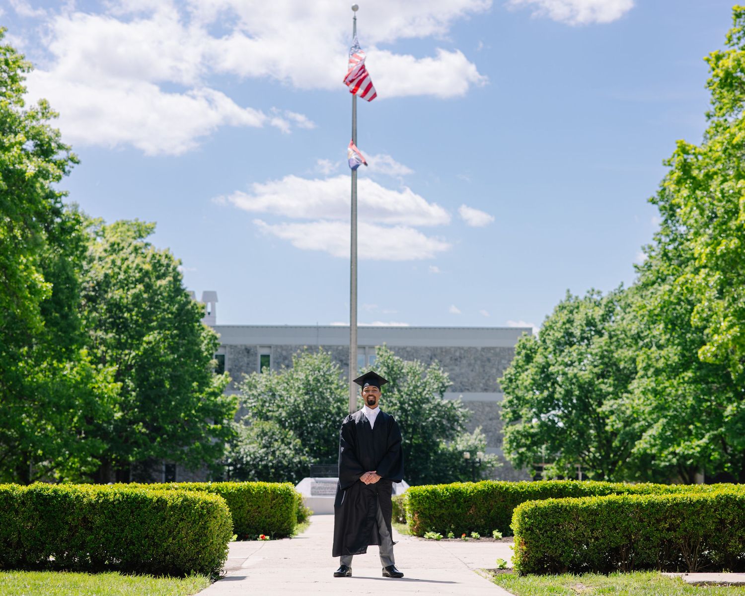 Graduate in cap and gown stands before an American flag on a flagpole, surrounded by lush green trees.