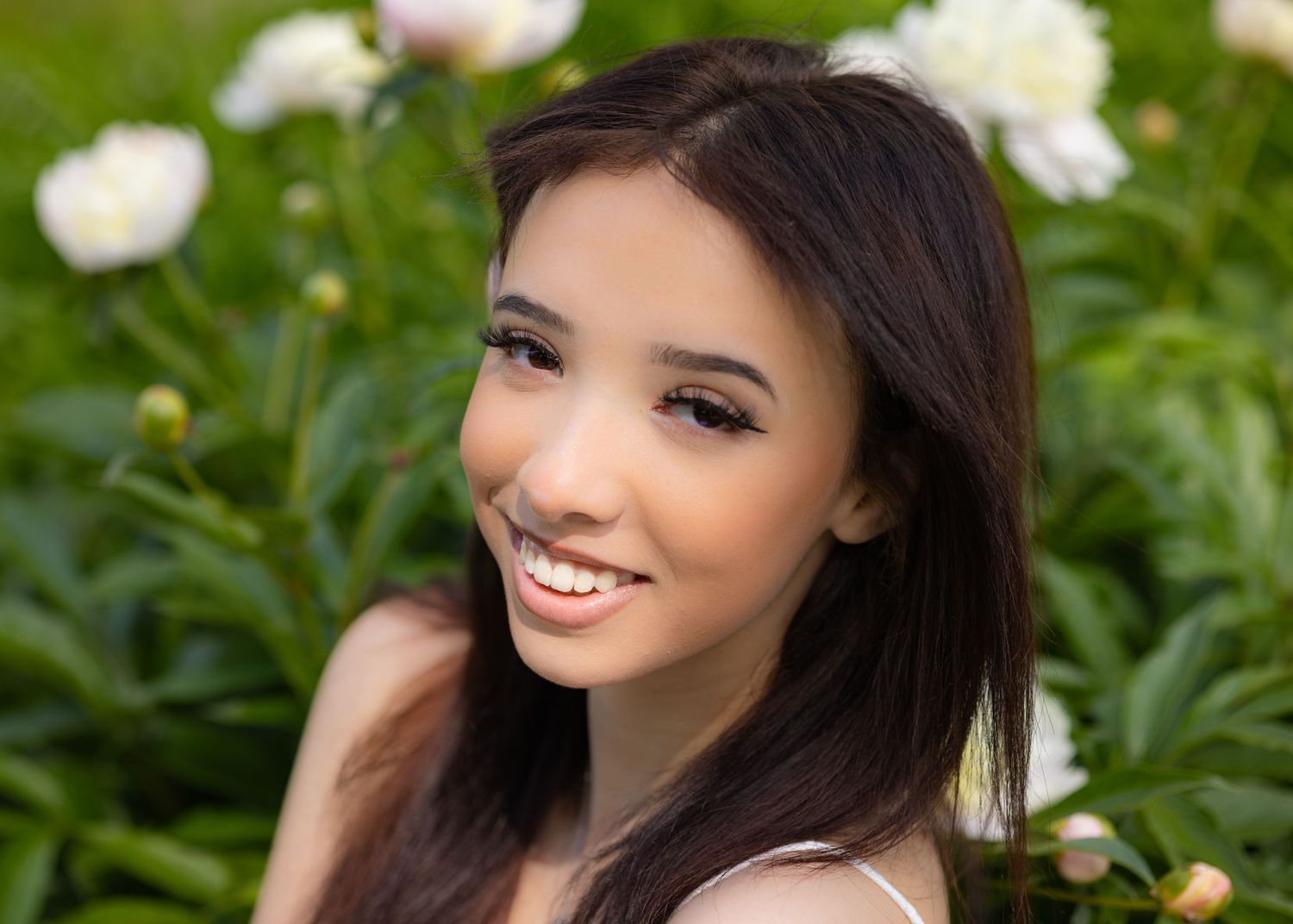 Smiling young woman with dark hair posing outdoors among white peony flowers in a lush green garden.