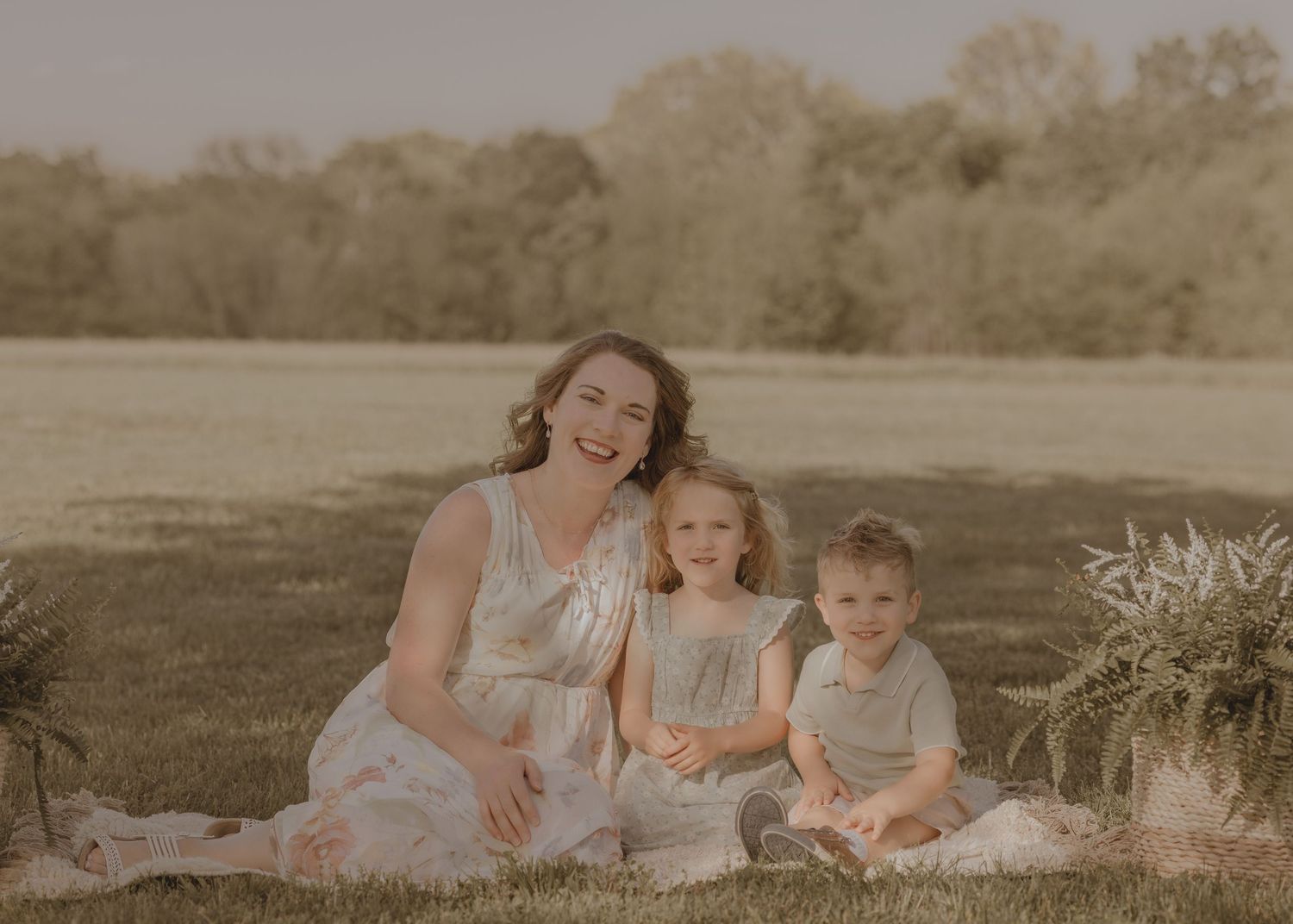 A smiling mother sits on a blanket in a sunny field with her two young children for an outdoor family portrait.