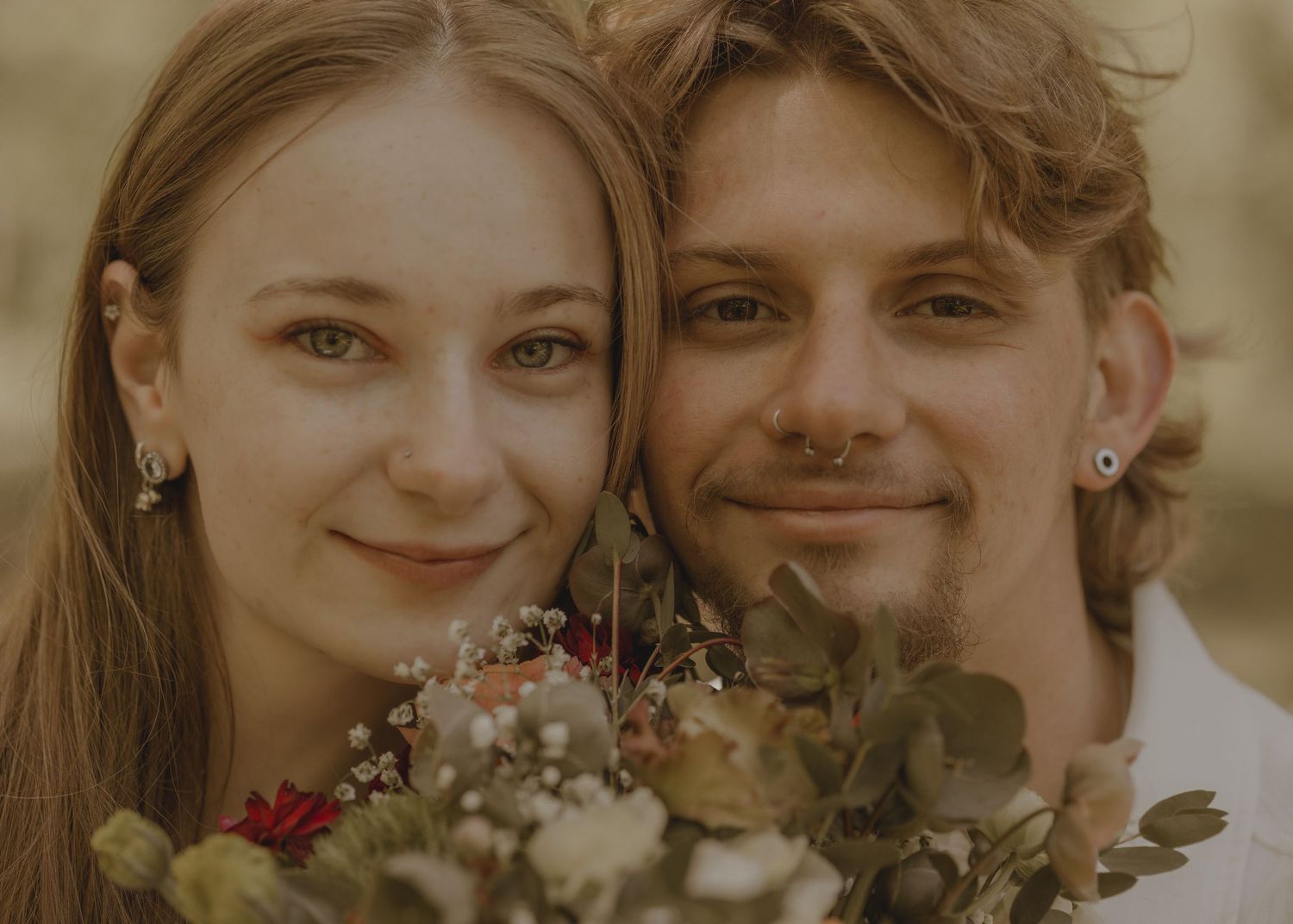 A smiling couple poses closely together holding a floral bouquet in a warm-toned portrait.