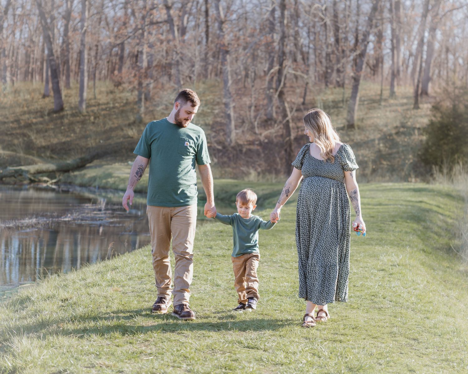 Family walking hand in hand along a pond in a scenic wooded park setting.