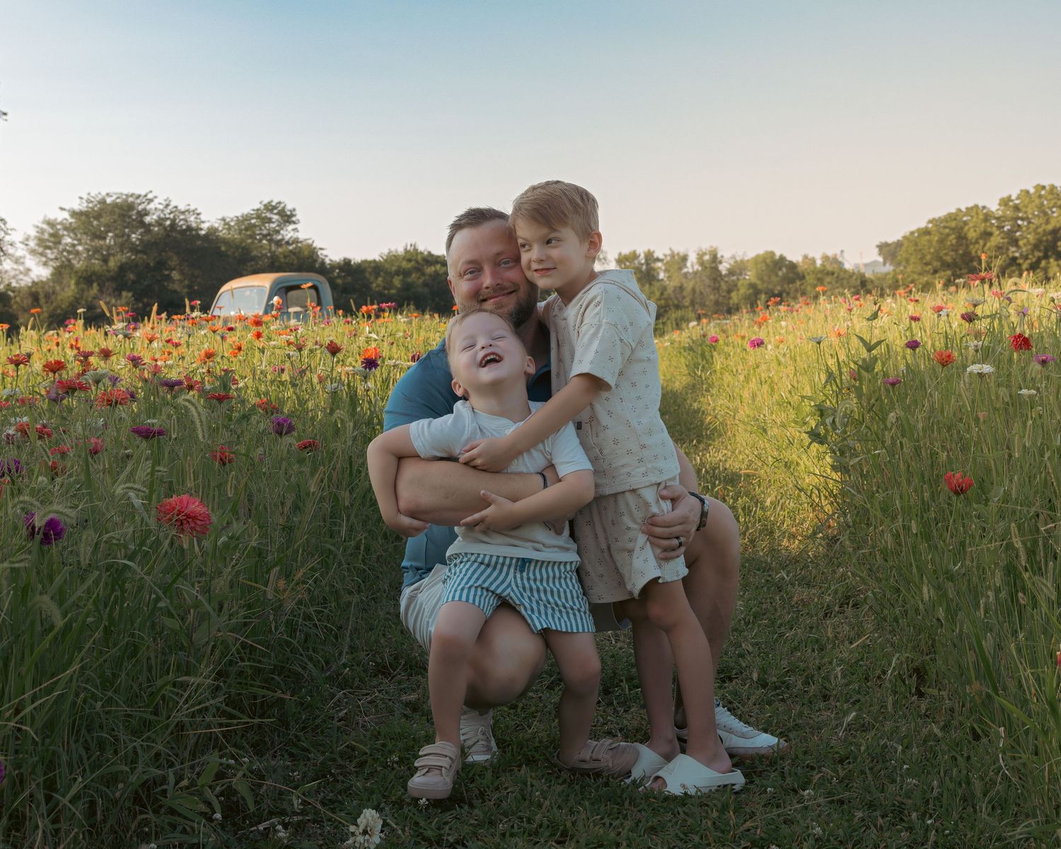 A father kneels in a vibrant wildflower field hugging his two young children, smiling warmly at golden hour.