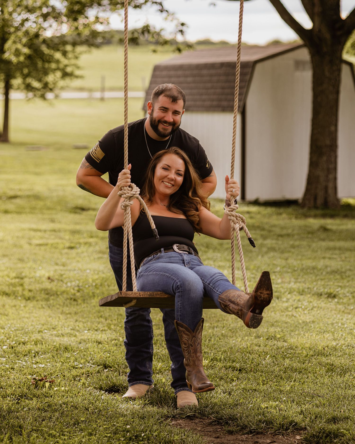 Couple posing on a wooden rope swing in a lush green farm yard, woman seated wearing cowboy boots.