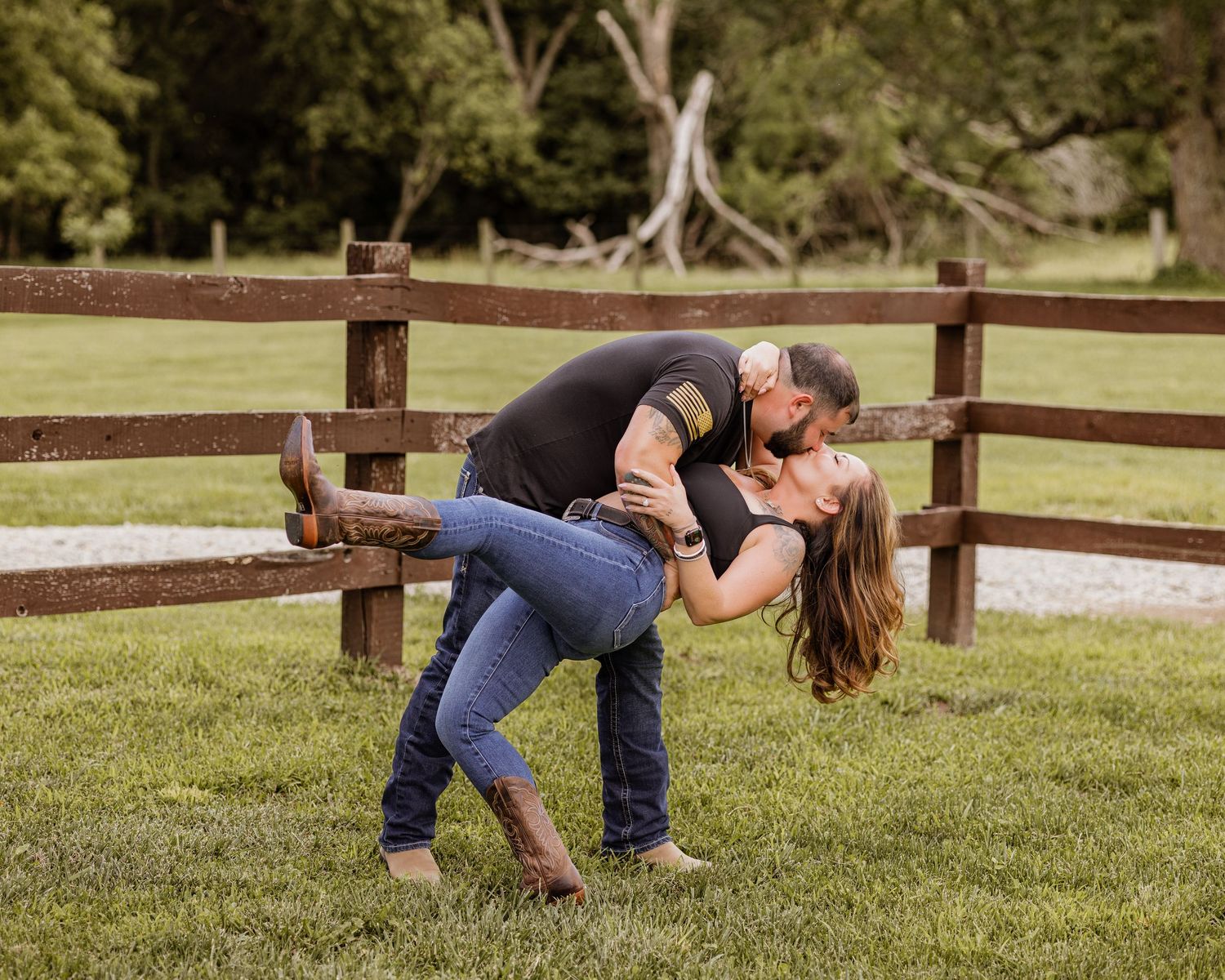 Romantic couple sharing a dip kiss near a rustic wooden fence on a green grassy farm.