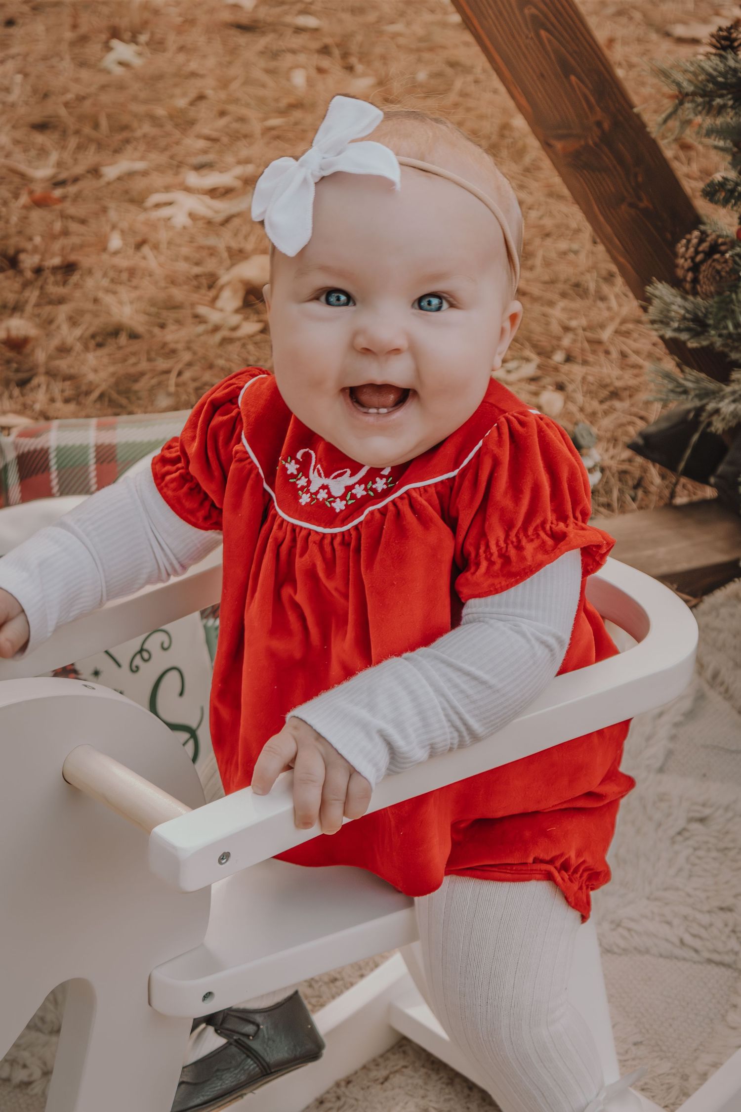 A baby in a red dress and white bow sits in a white wooden rocking chair during an outdoor photo session.