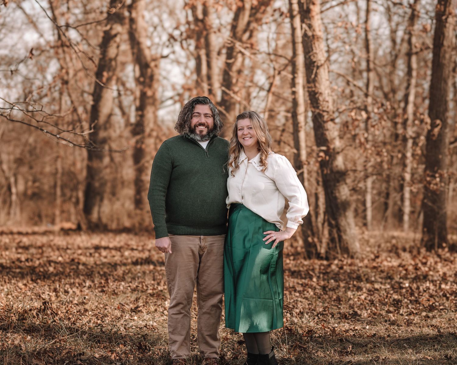 A couple stands together in an autumn forest setting with warm brown tones and fallen leaves on the ground.