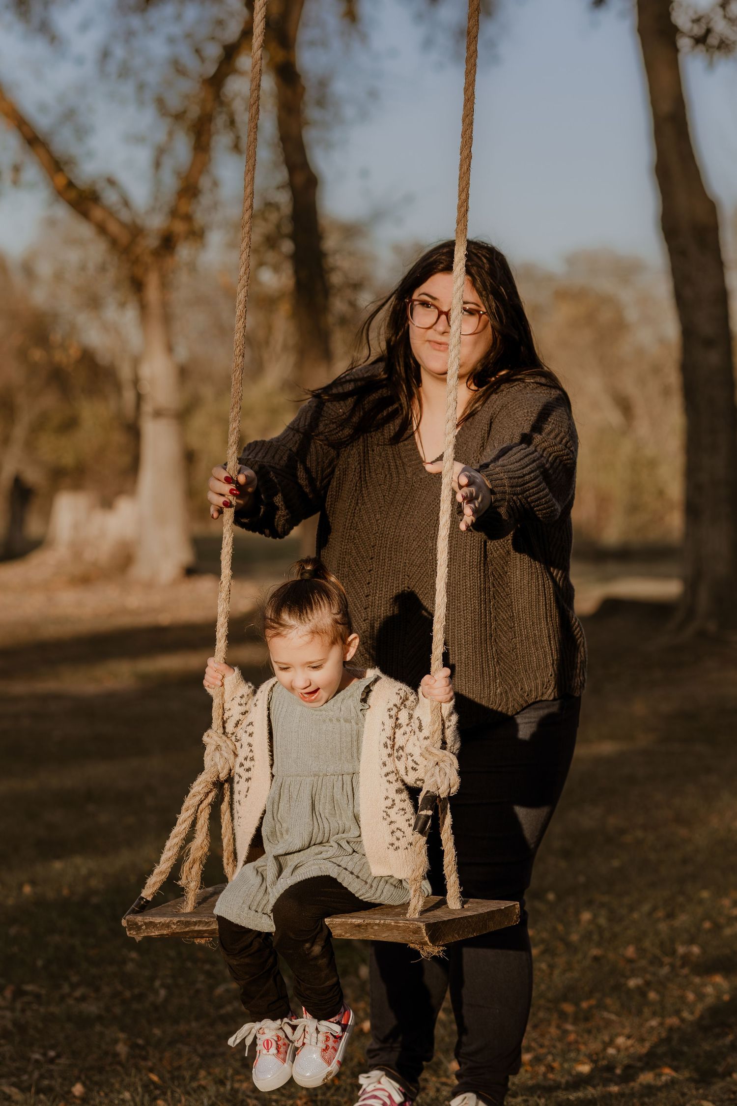 A heartwarming scene of a child enjoying a wooden rope swing while being pushed in a wooded setting at sunset.