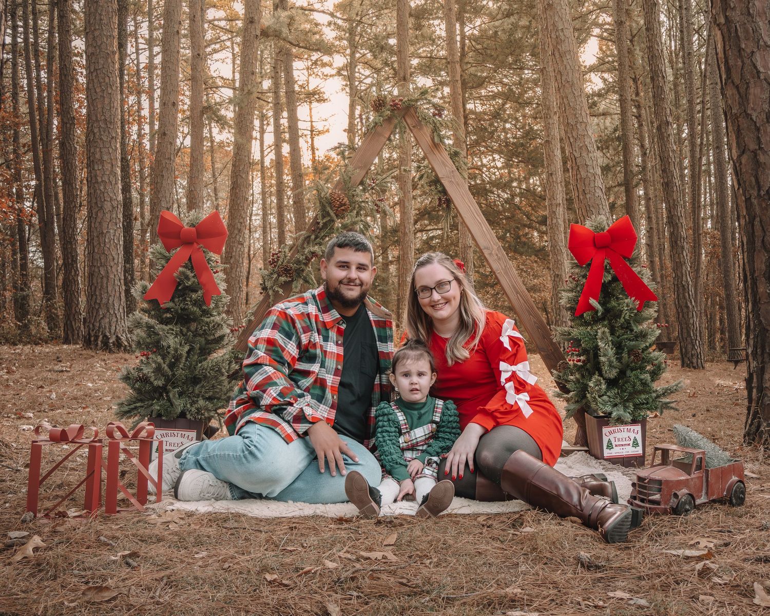 A family sits together in a pine forest during golden hour for holiday photos with red Christmas decorations.