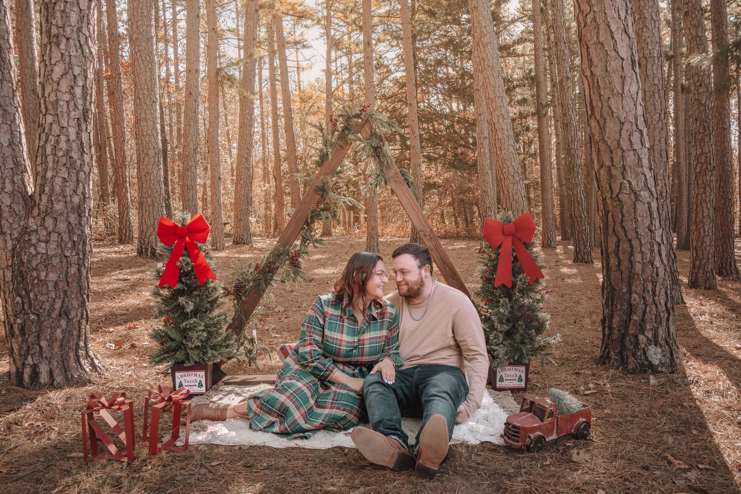 A couple sits together in a forest setting decorated with red bows and holiday decor during a winter photo session.