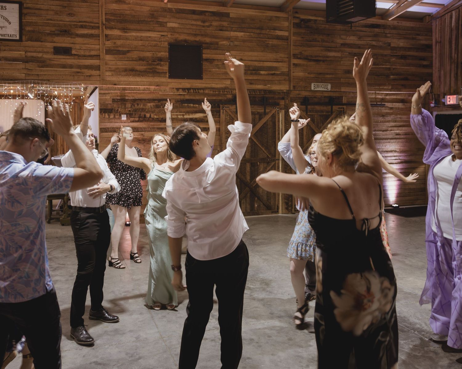 Wedding guests dance with arms raised at a rustic barn reception celebration.