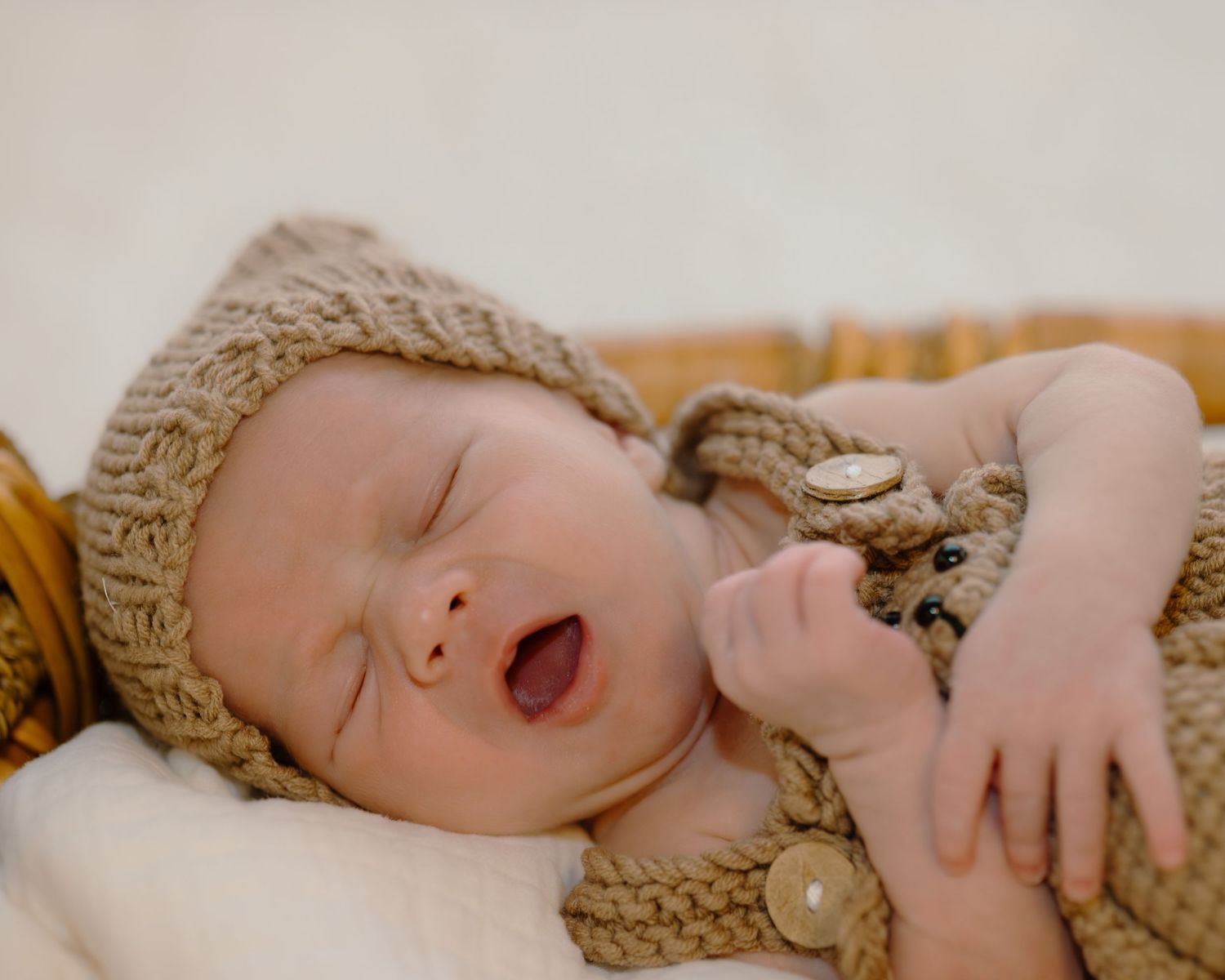 Sleeping newborn baby in knit bear outfit and hat, yawning with tiny hand raised in cozy basket.