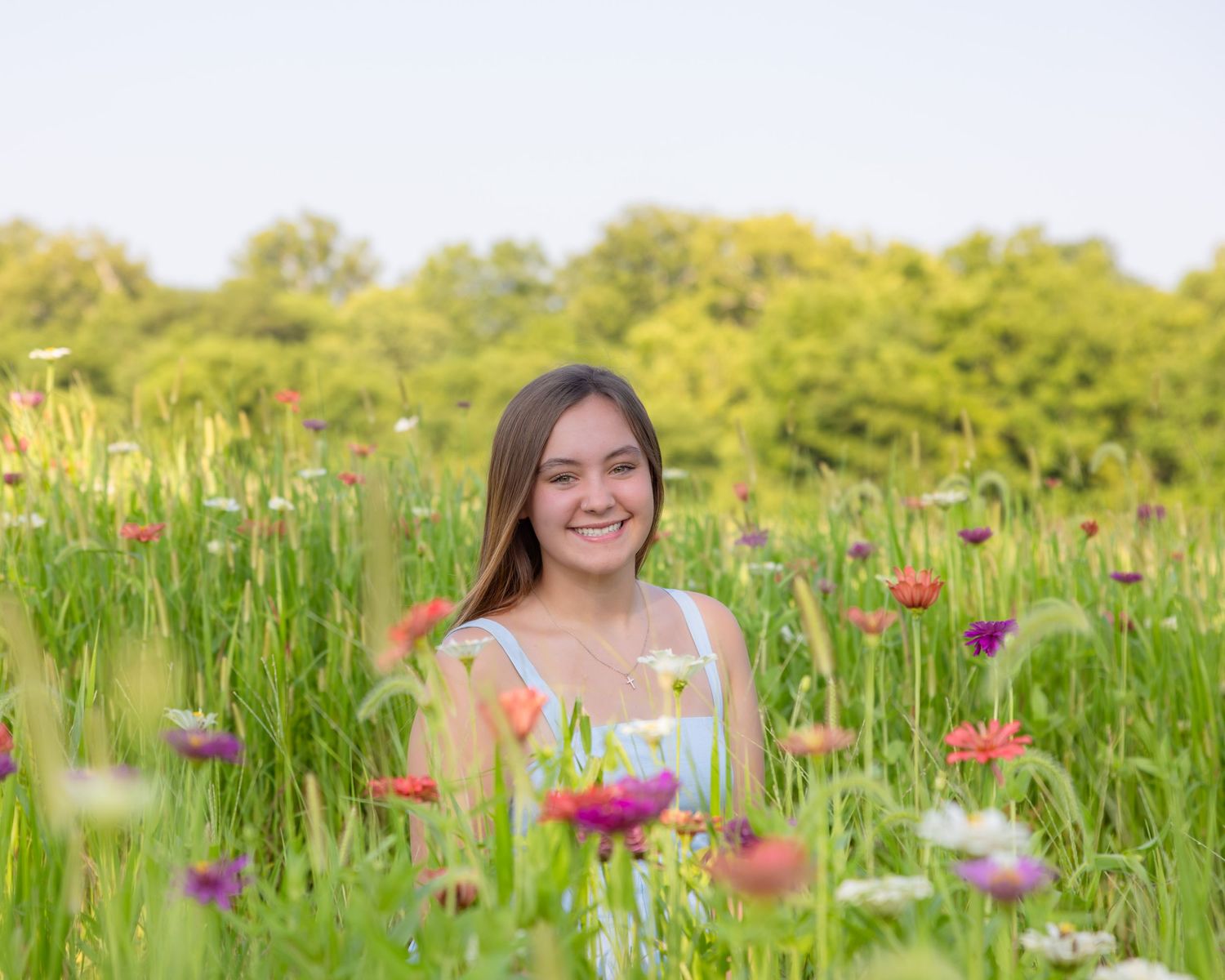 Smiling young woman sitting in a vibrant wildflower meadow with colorful blooms and lush green trees.