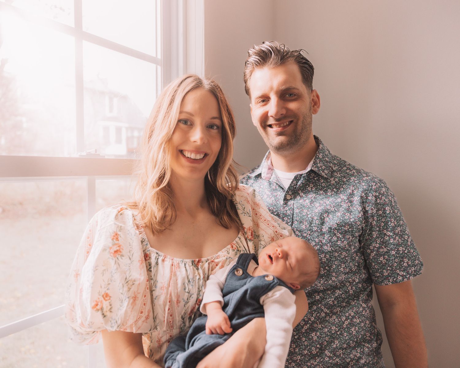 Smiling couple holding their newborn baby near a bright window in a warm, softly lit portrait.