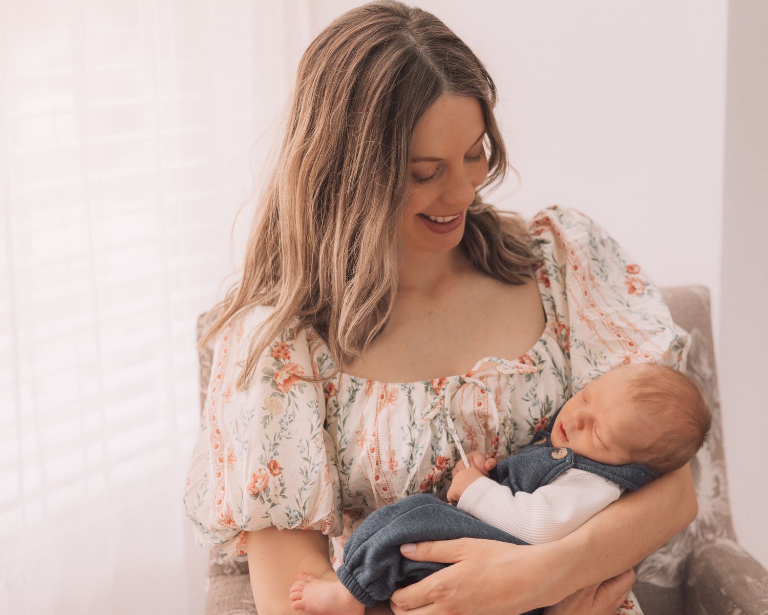 Smiling mother in floral dress lovingly gazing at her sleeping newborn baby in her arms.