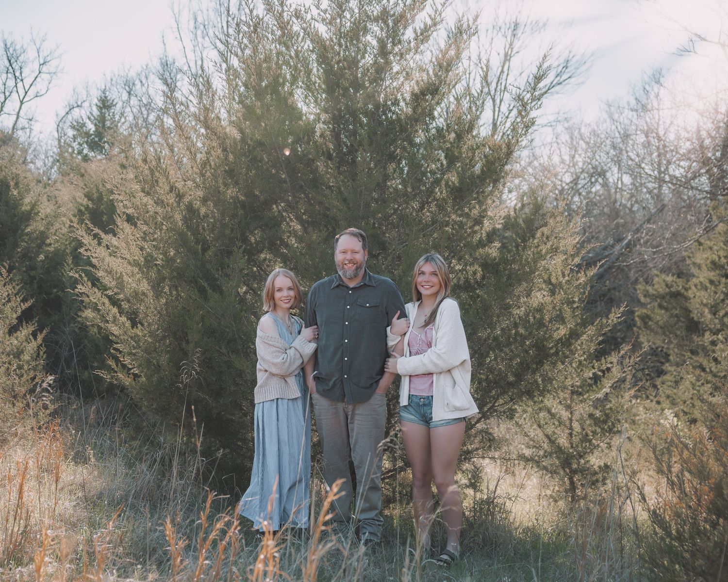 Family of three smiling outdoors among tall golden shrubs at sunset.