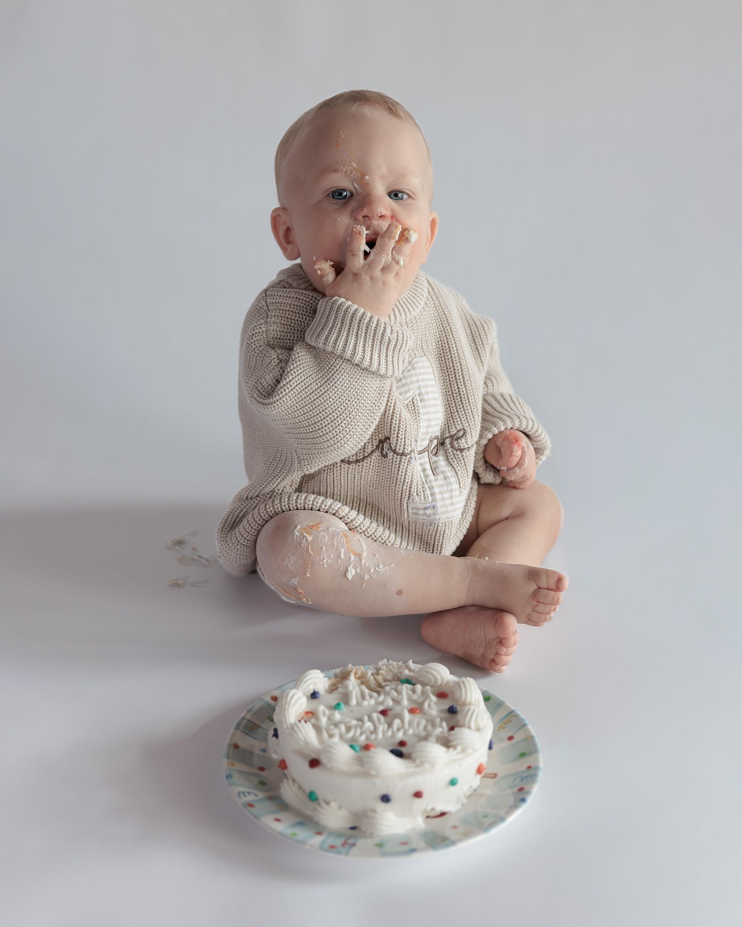 Adorable baby in cream knit romper eats birthday cake with hands during messy first birthday smash cake session.