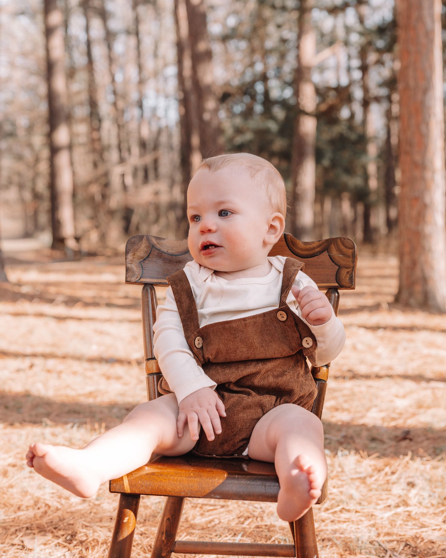 Baby wearing brown overalls sits in wooden chair during autumn photoshoot.