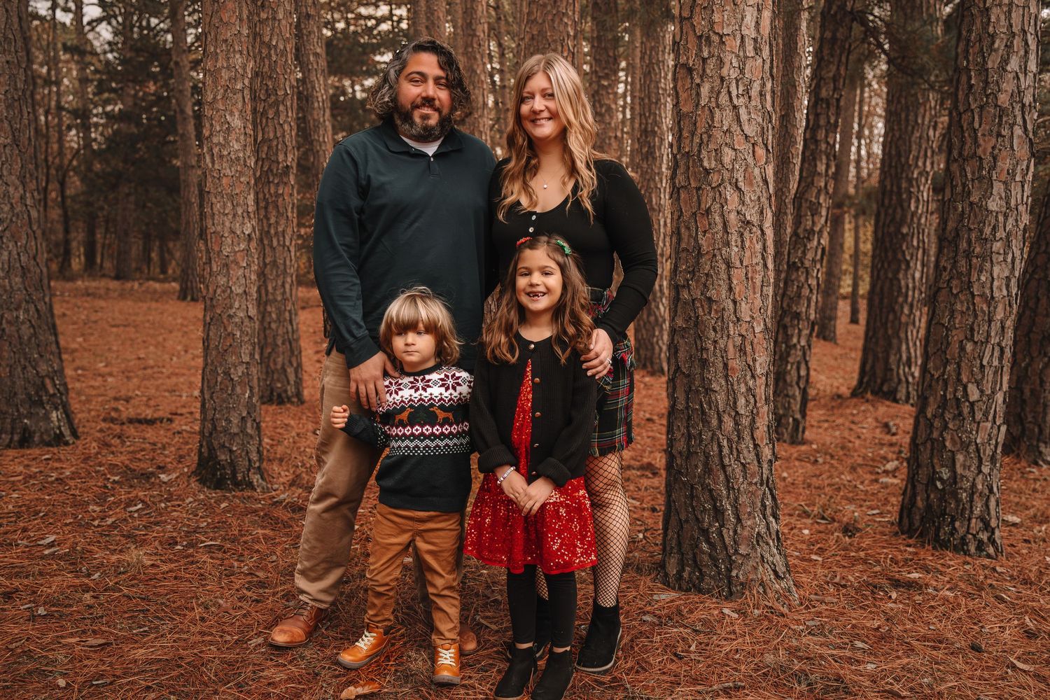 Family standing together in a rustic autumn forest setting with tall pine trees and orange fallen leaves on the ground.