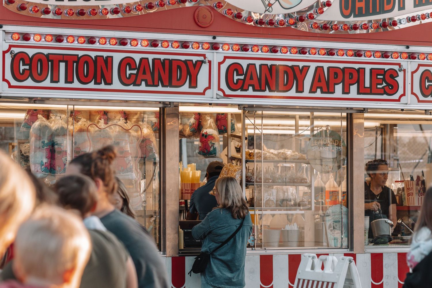 Customers lined up at a carnival concession stand selling cotton candy and candy apples.
