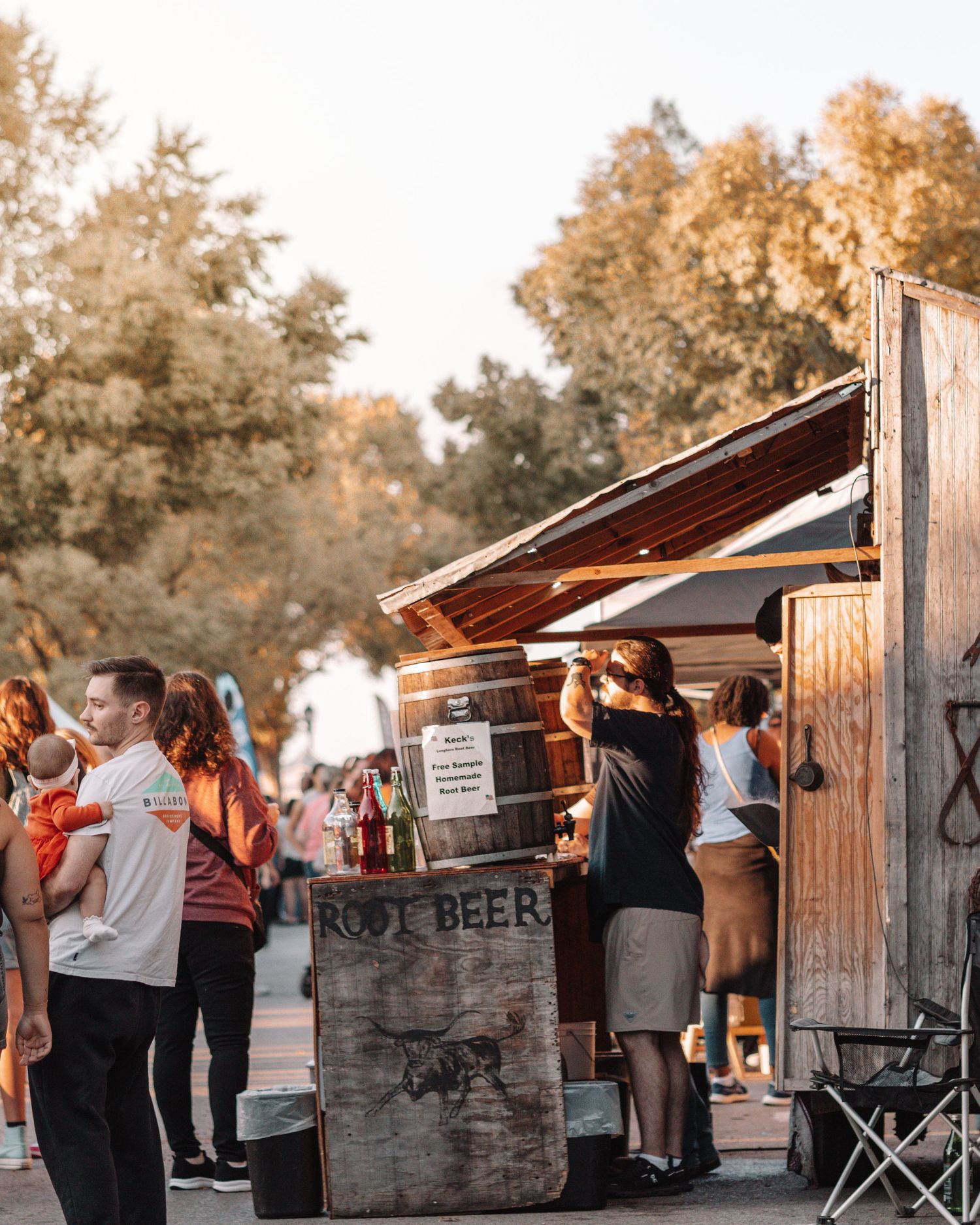 People in casual attire gathered at wooden food vendor stalls during sunset at an outdoor event.