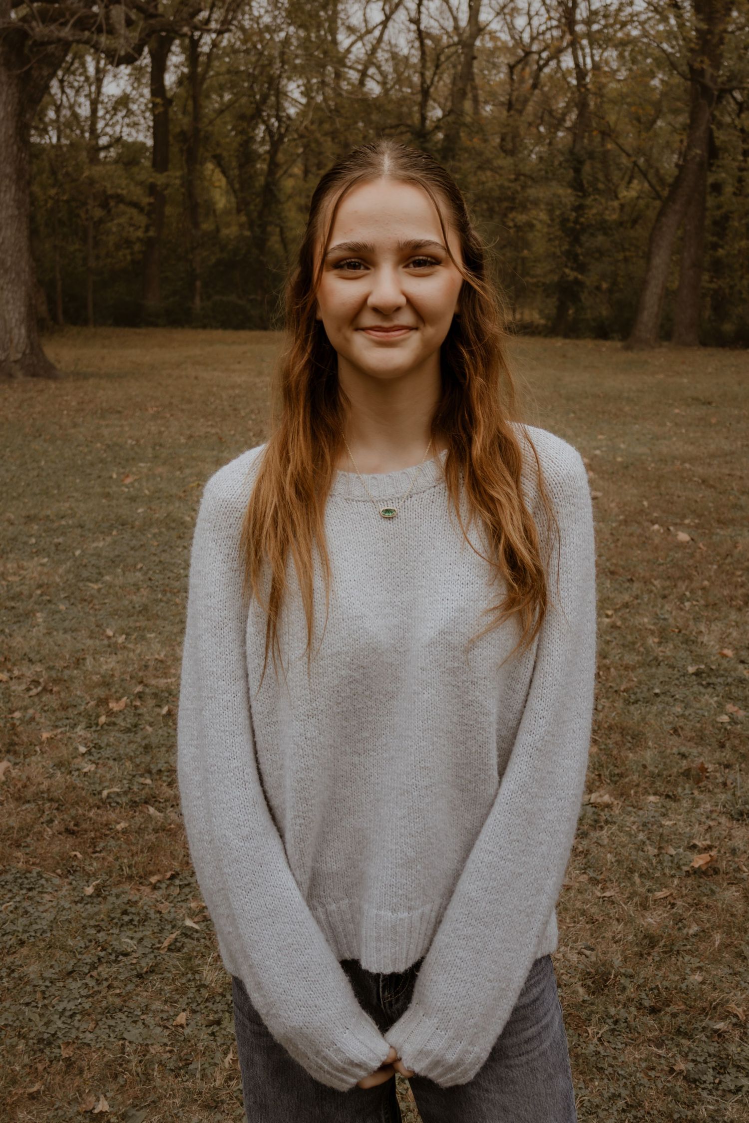 A person in a gray sweater poses outdoors in a wooded area during autumn.