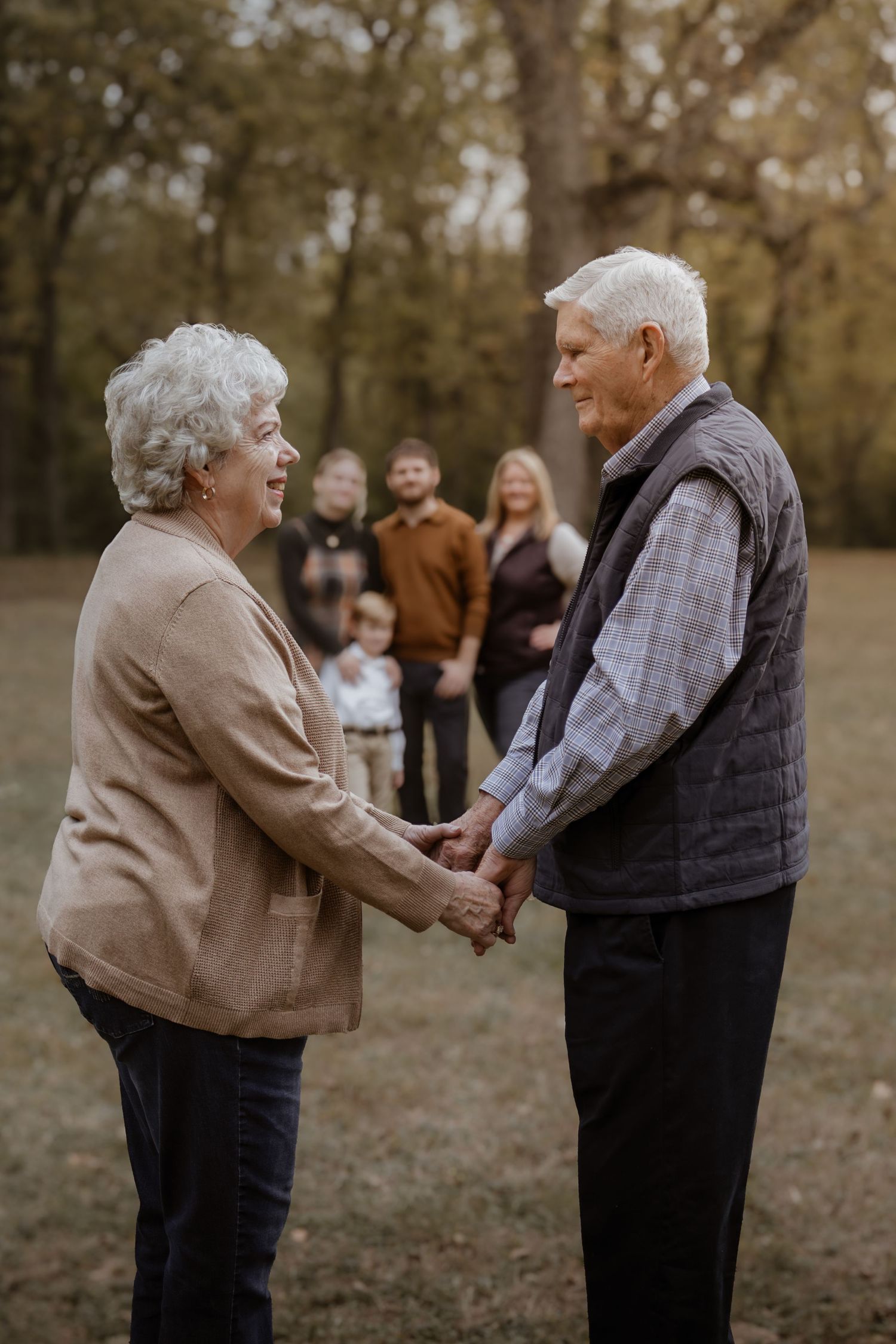 An elderly couple holds hands tenderly in a wooded outdoor setting as others look on in the background.