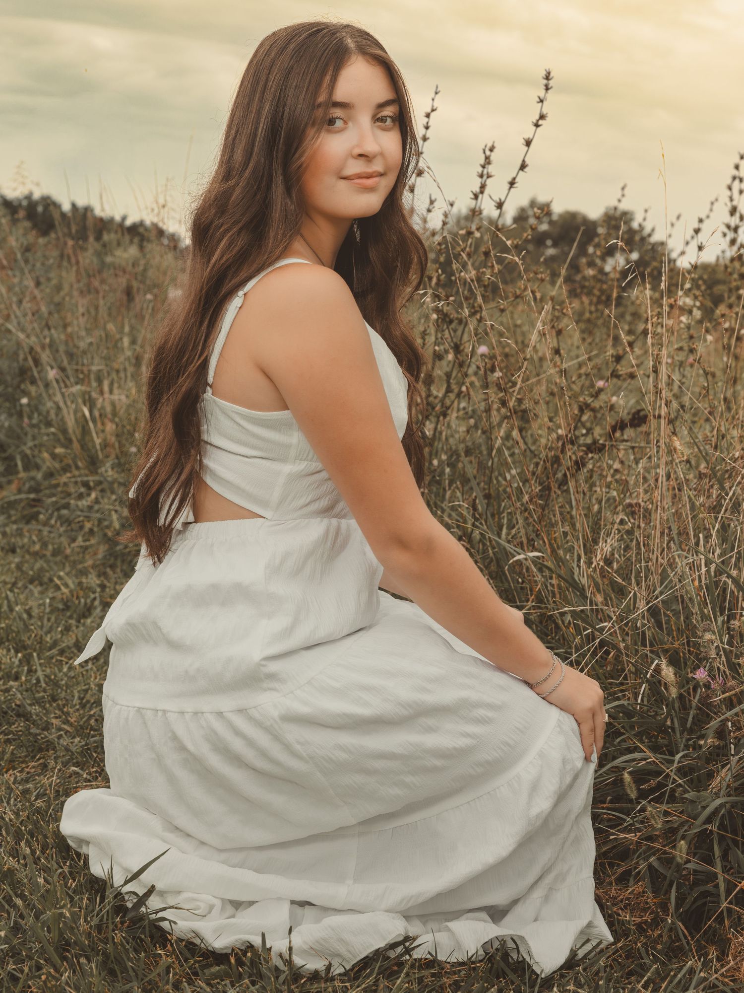 Brunette woman in white cutout dress sitting in golden grass field.