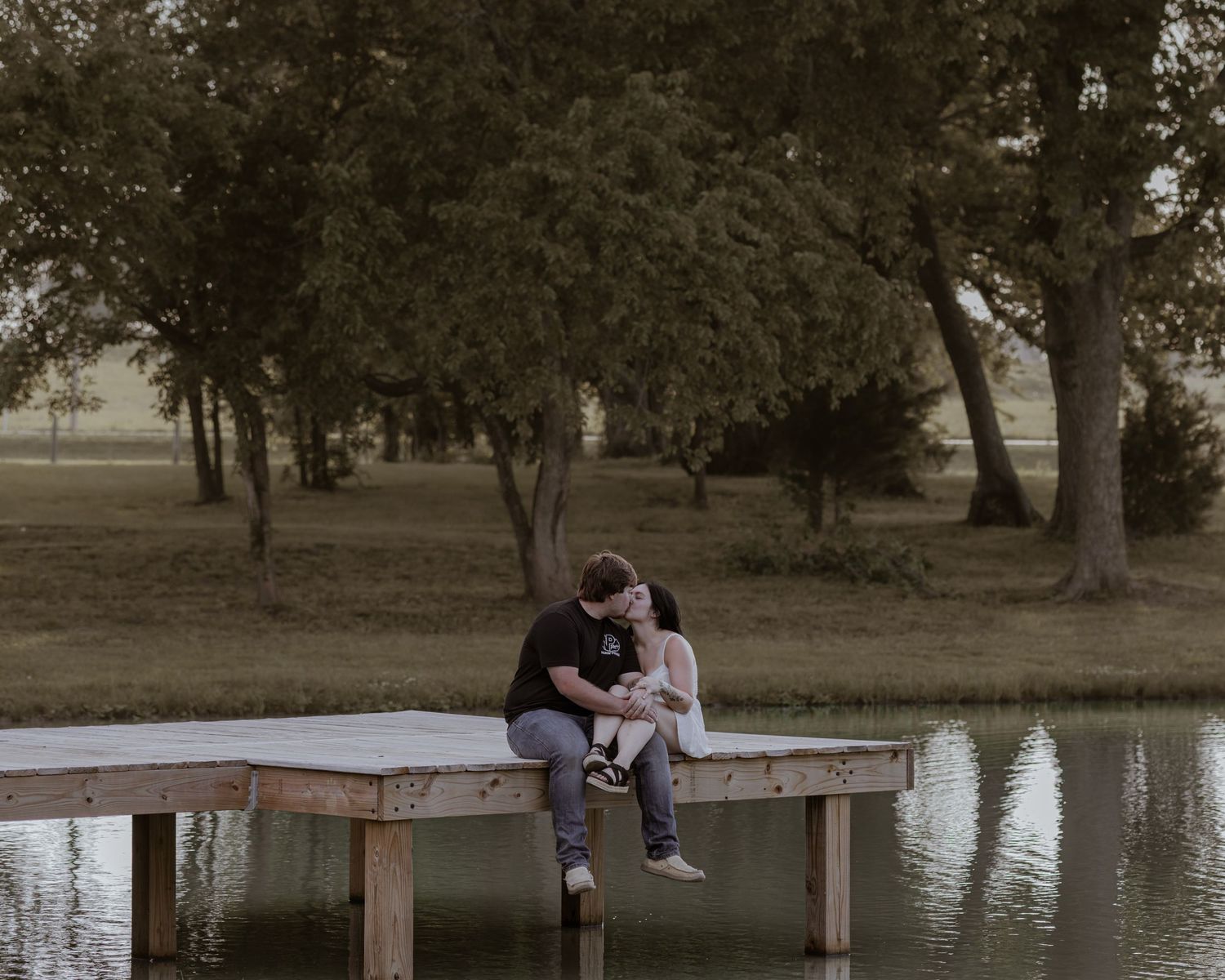 A couple shares a kiss while sitting on a wooden dock over a calm pond surrounded by lush green trees.
