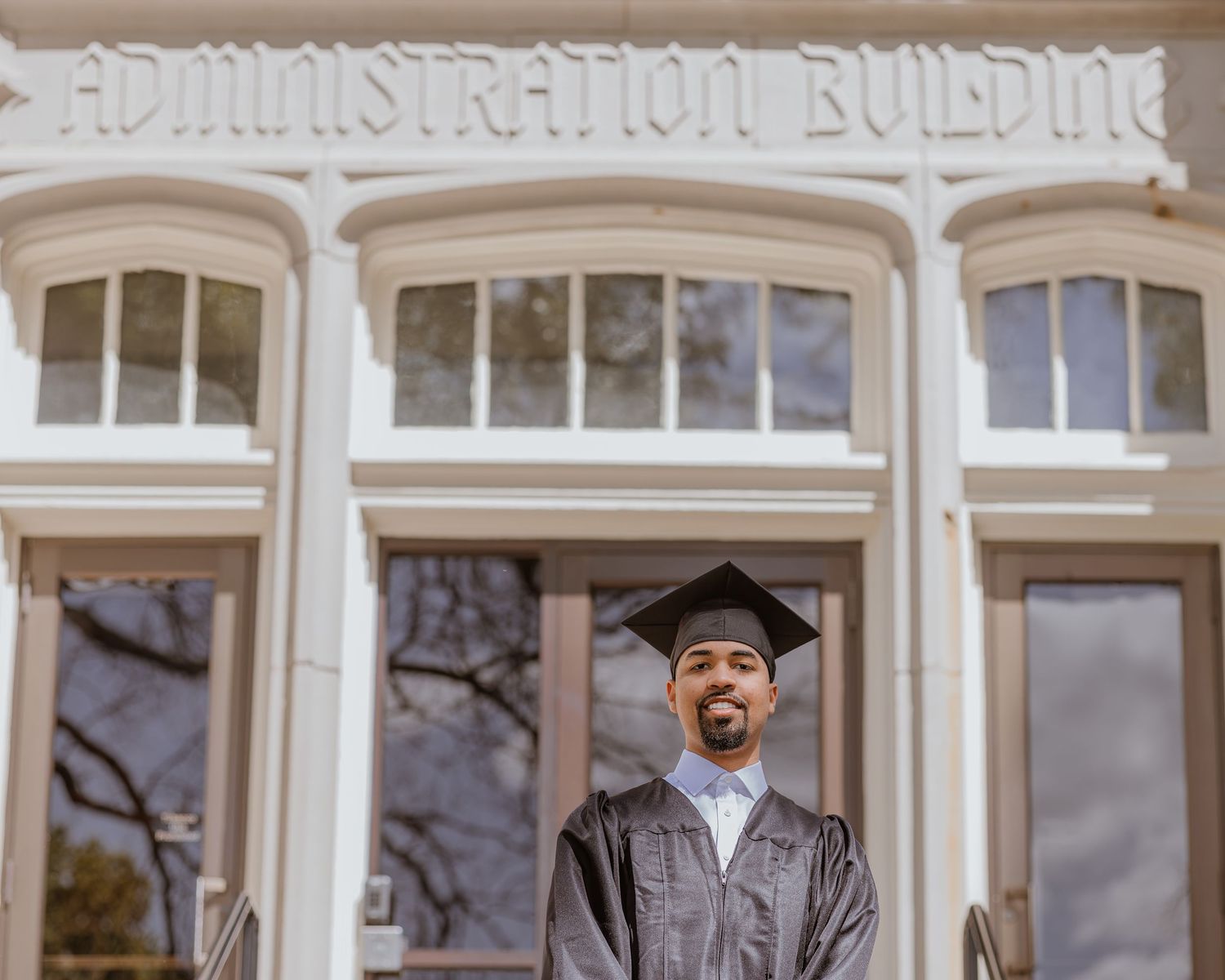 Graduate in cap and gown stands proudly before a historic Administration Building on graduation day.