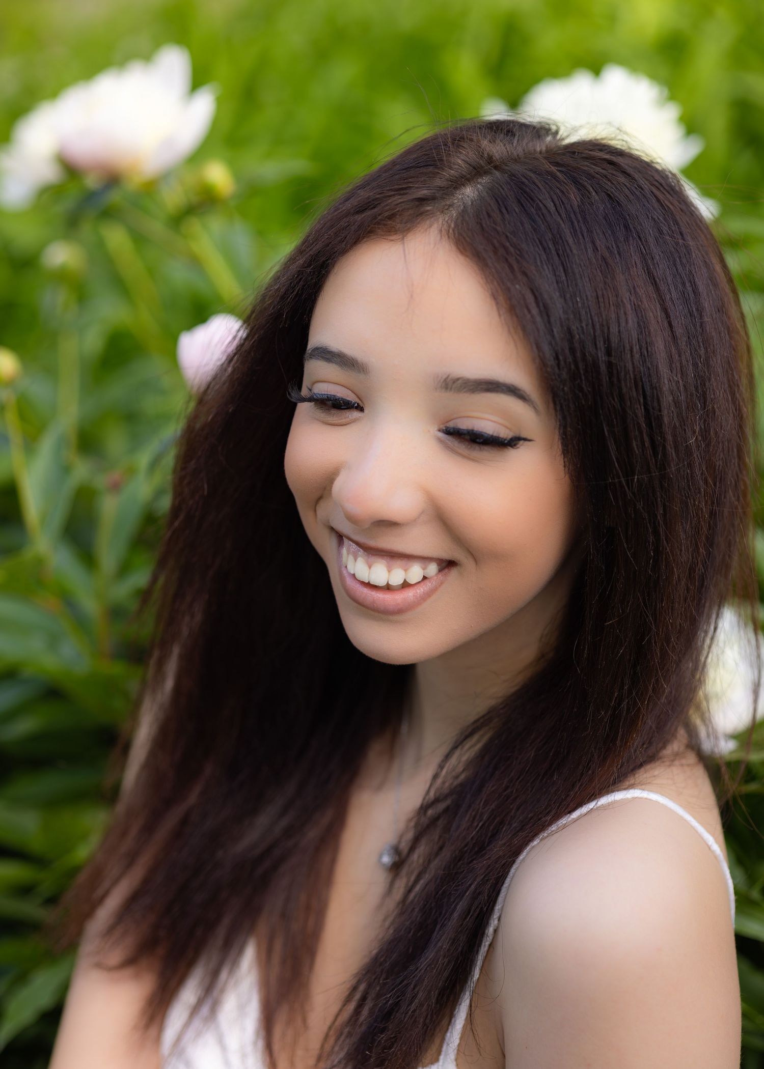 Beautiful young woman with long dark hair smiling among white peony flowers in a garden.