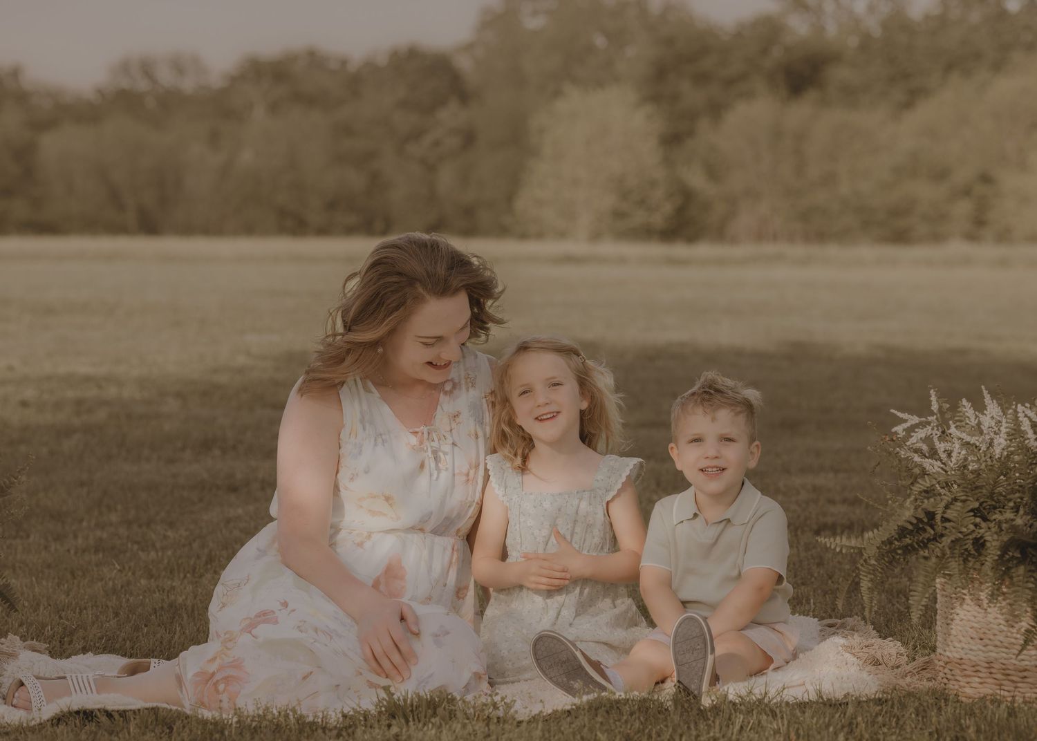 A mother sits on a blanket in a sunny field smiling at her two young children in coordinating outfits.