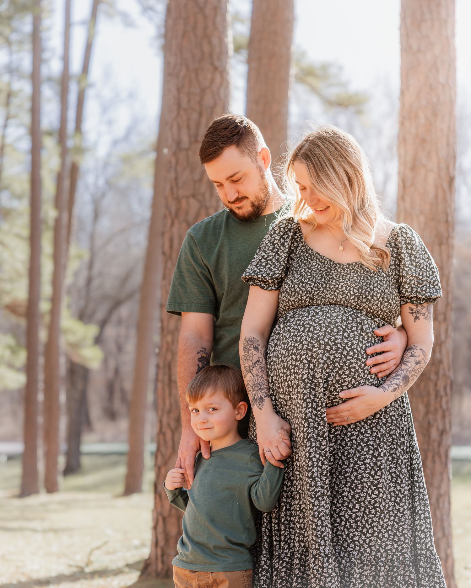 Family maternity photo in a pine forest with matching green outfits and floral dress.