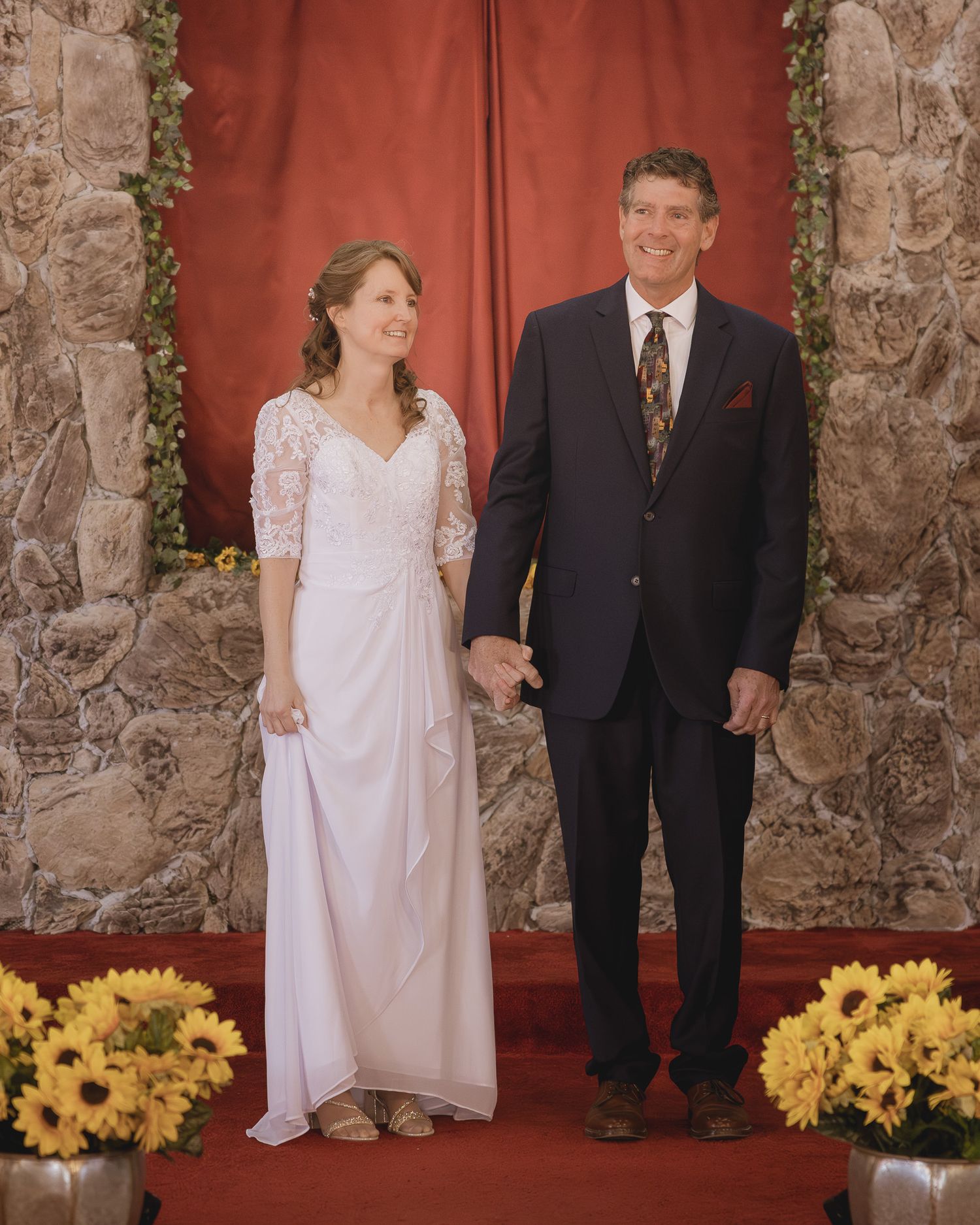 A bride in a white lace dress and groom in dark suit stand on red carpet with yellow flowers decorating stone archway.