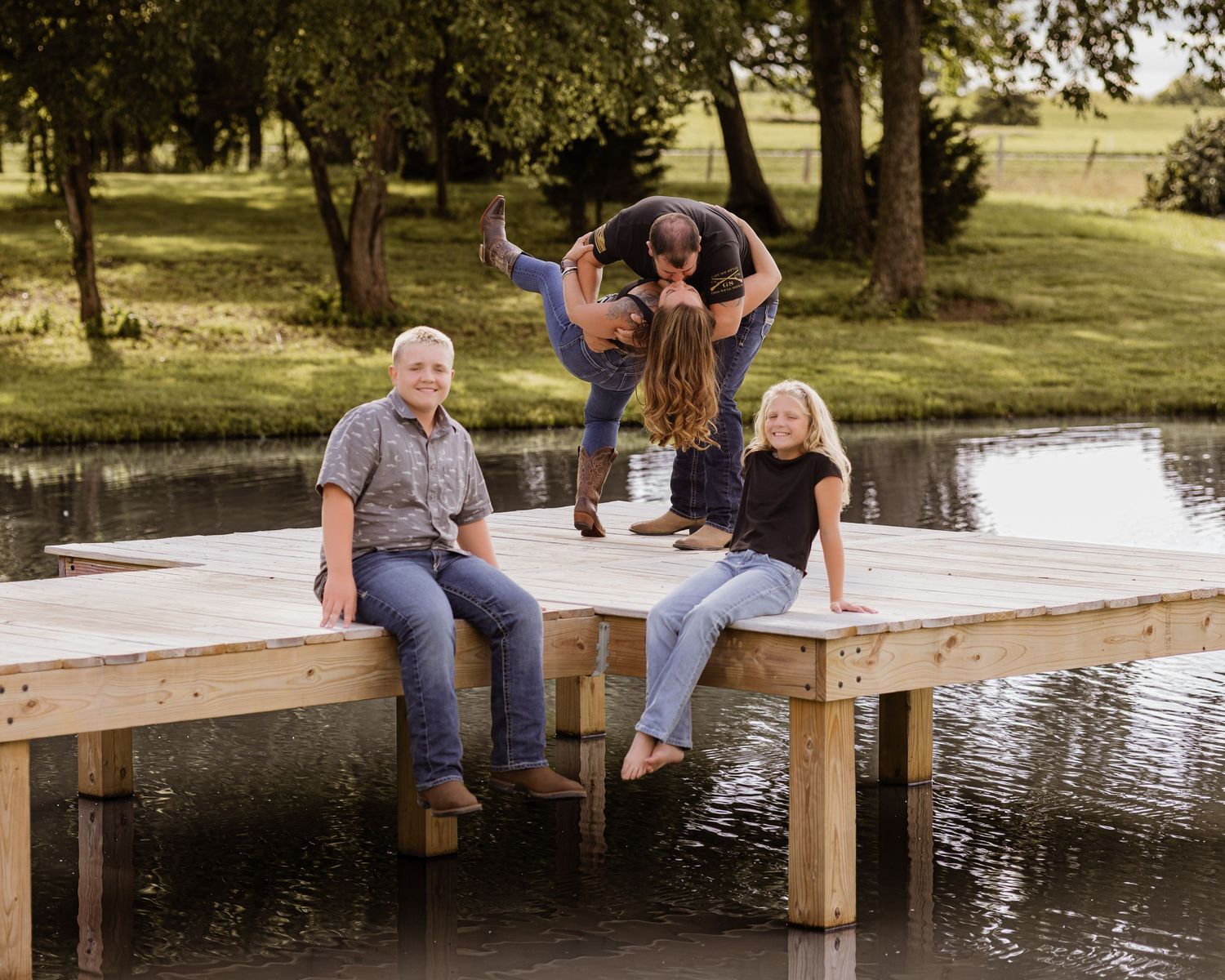 Family of four enjoying a sunny day on a wooden dock by a pond, with two playfully lifting a child in the air.