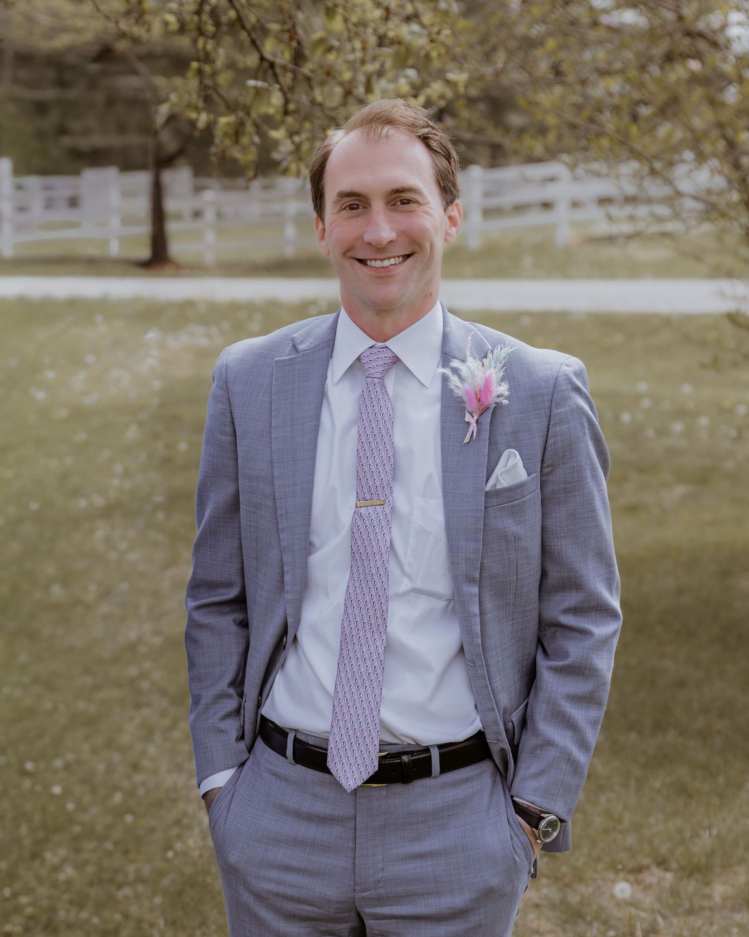 Smiling groom in a gray suit with pink tie and boutonniere standing outdoors on wedding day.
