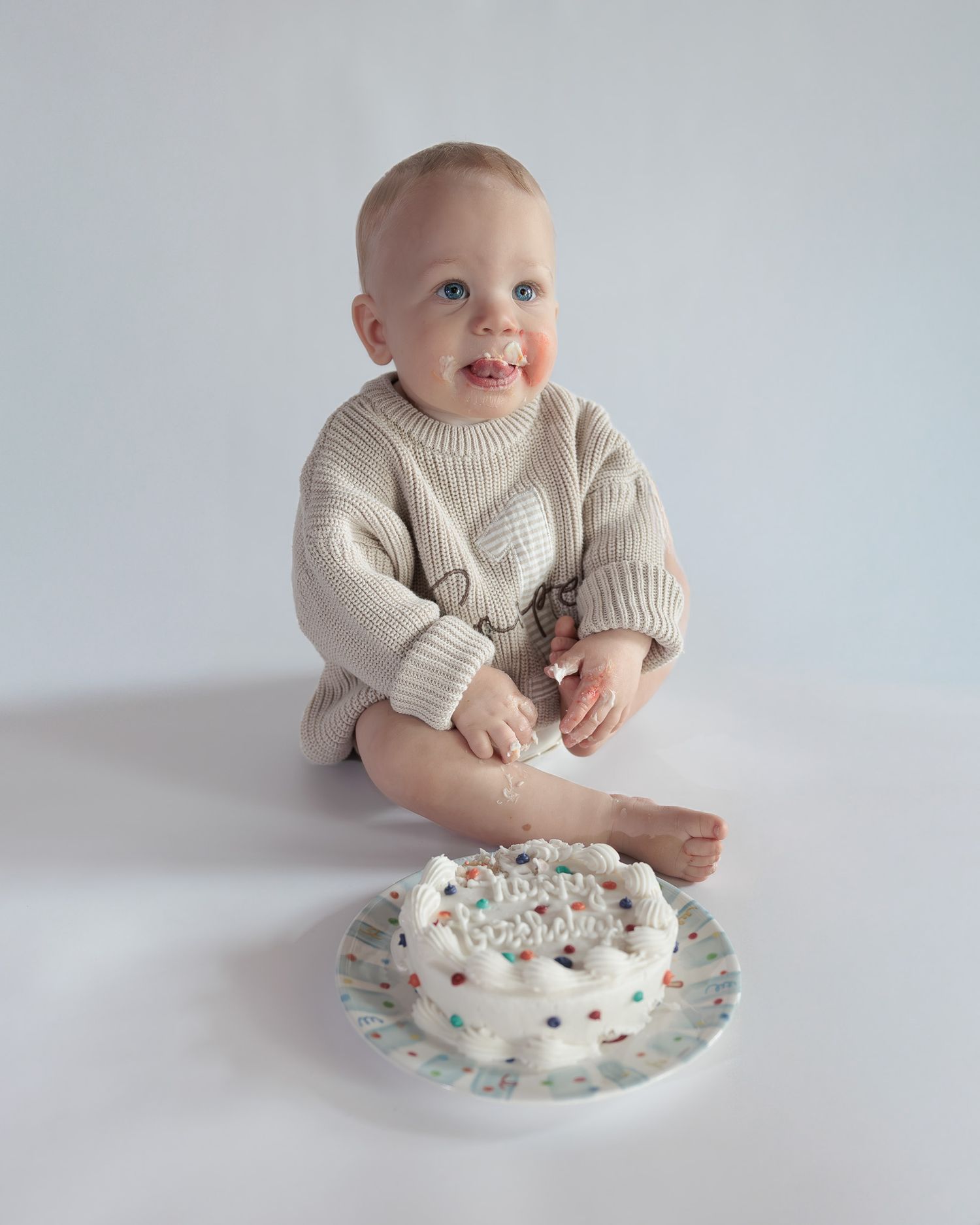 Cute baby in cream knit sweater romper sits beside a small white birthday smash cake on white background.