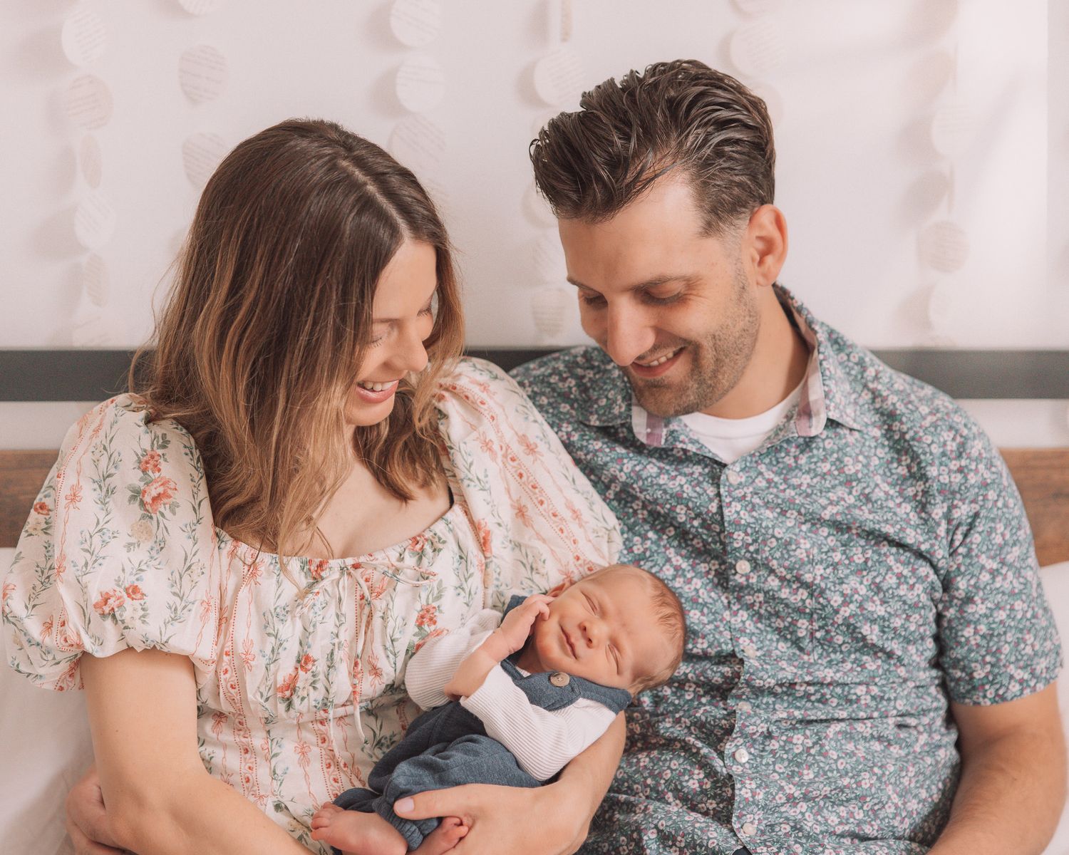 Smiling parents sitting on a bed, gazing adoringly at their sleeping newborn baby in soft lighting.
