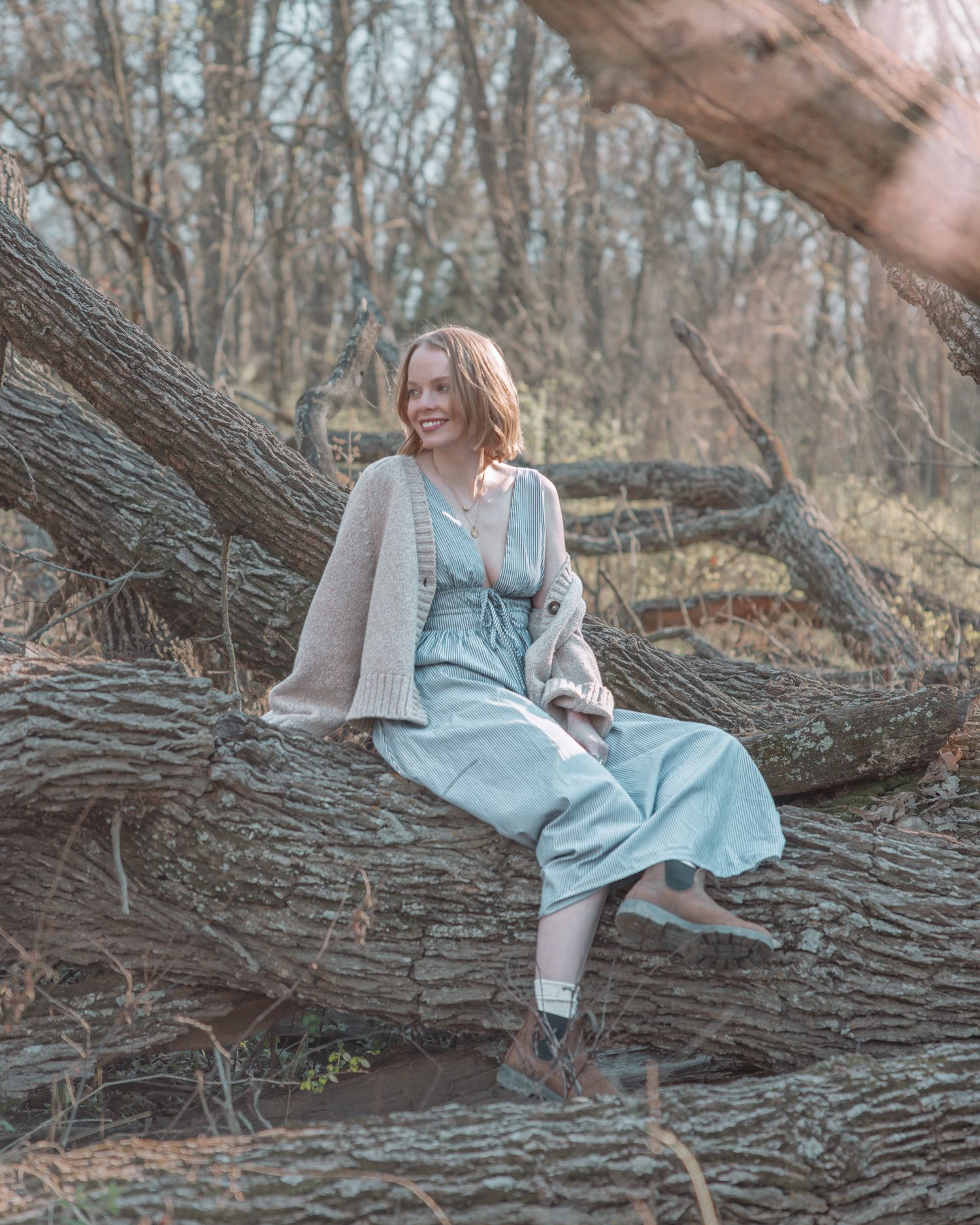 Smiling young woman in light blue dress and beige cardigan sitting on a fallen tree in a bare winter forest.